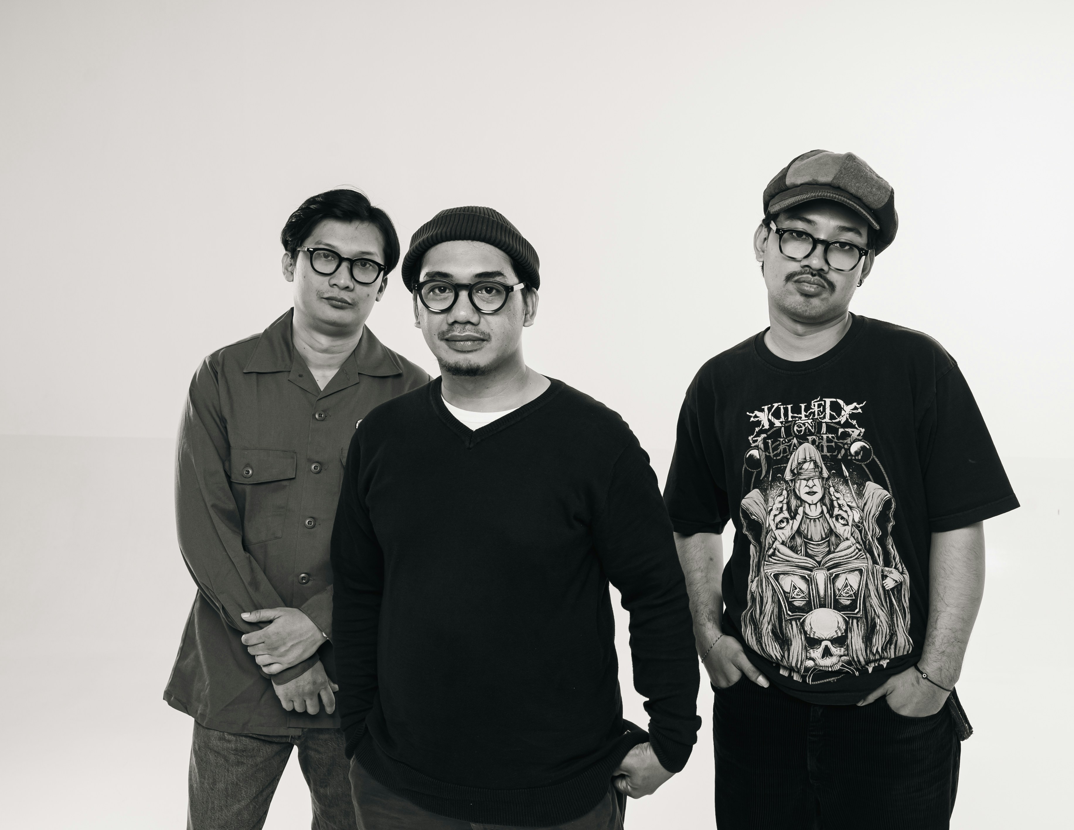 Three men stand close together, smiling faintly. All wear glasses and casual, dark clothing. One sports a cap, another a beanie, the third a buttoned shirt. They appear relaxed, slightly amused