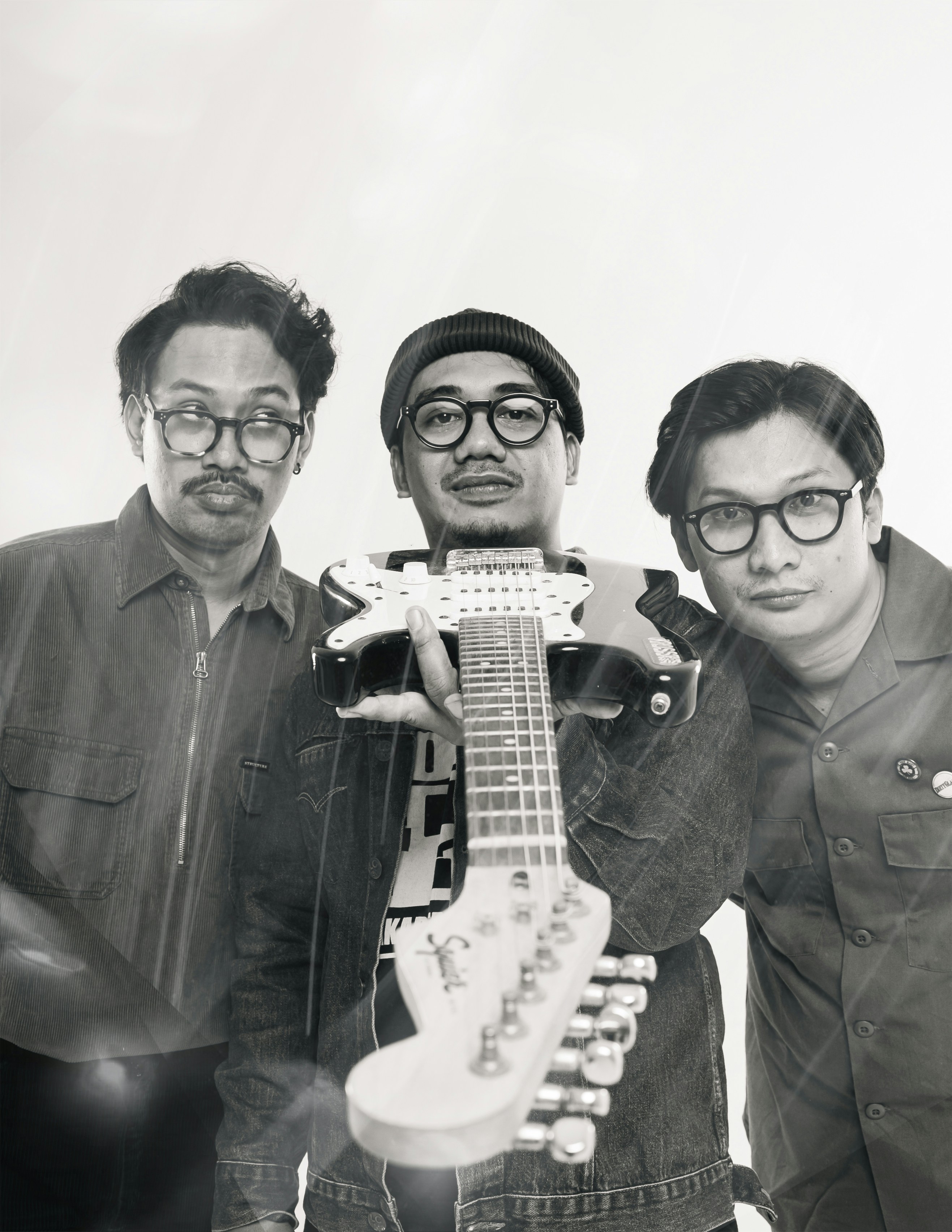 Three men stand close together, smiling faintly. All wear glasses and casual, dark clothing. One sports a cap, another a beanie, the third a buttoned shirt. They appear relaxed, slightly amused