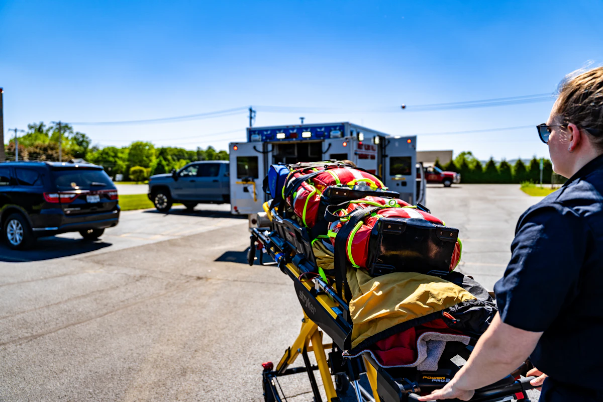 Une ambulancière pousse un brancard chargé de sacs de matériel vers une ambulance aux portes arrière ouvertes, sur un parking ensoleillé