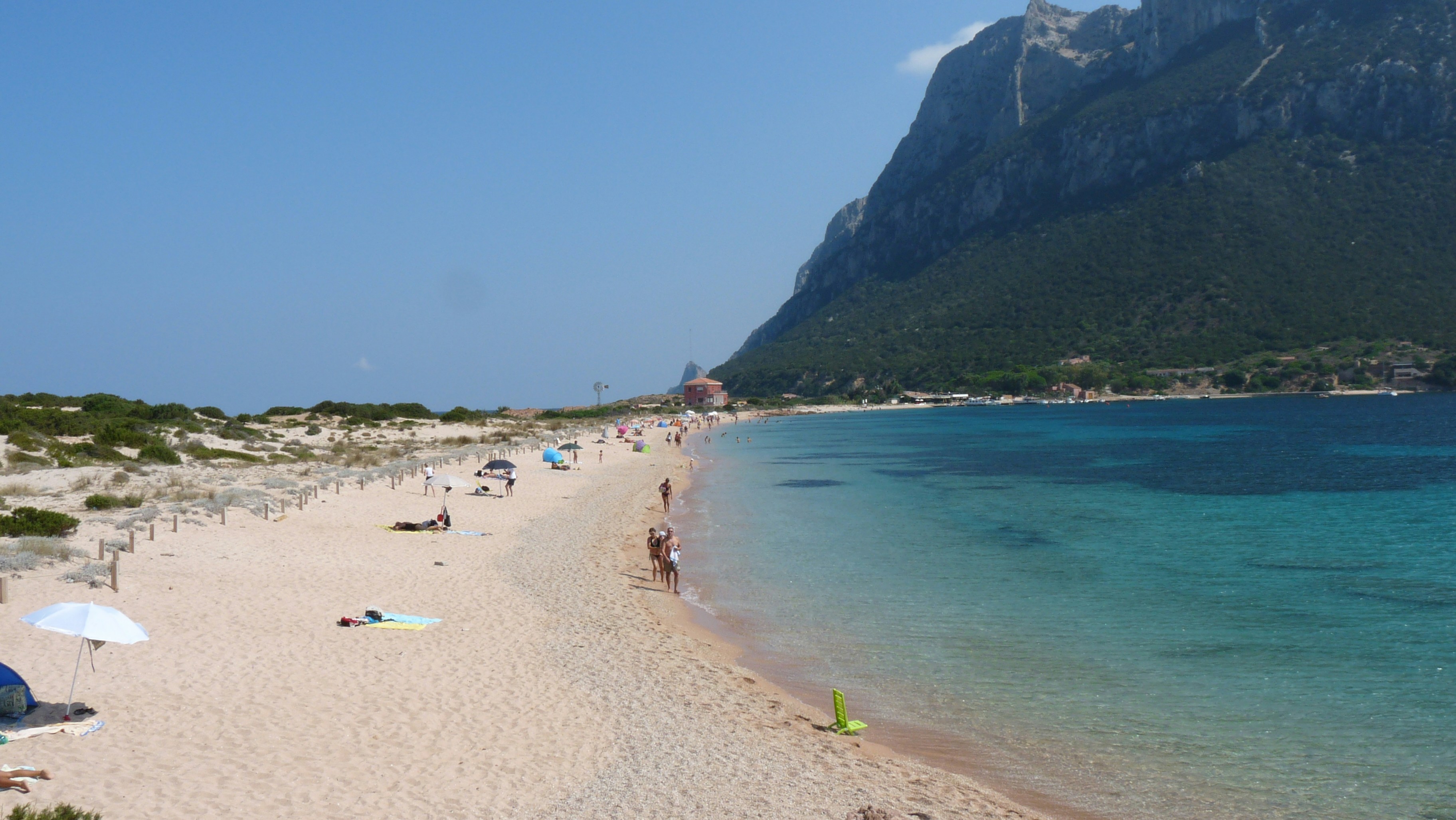 Sandy beach with people, clear blue water, and a mountain.