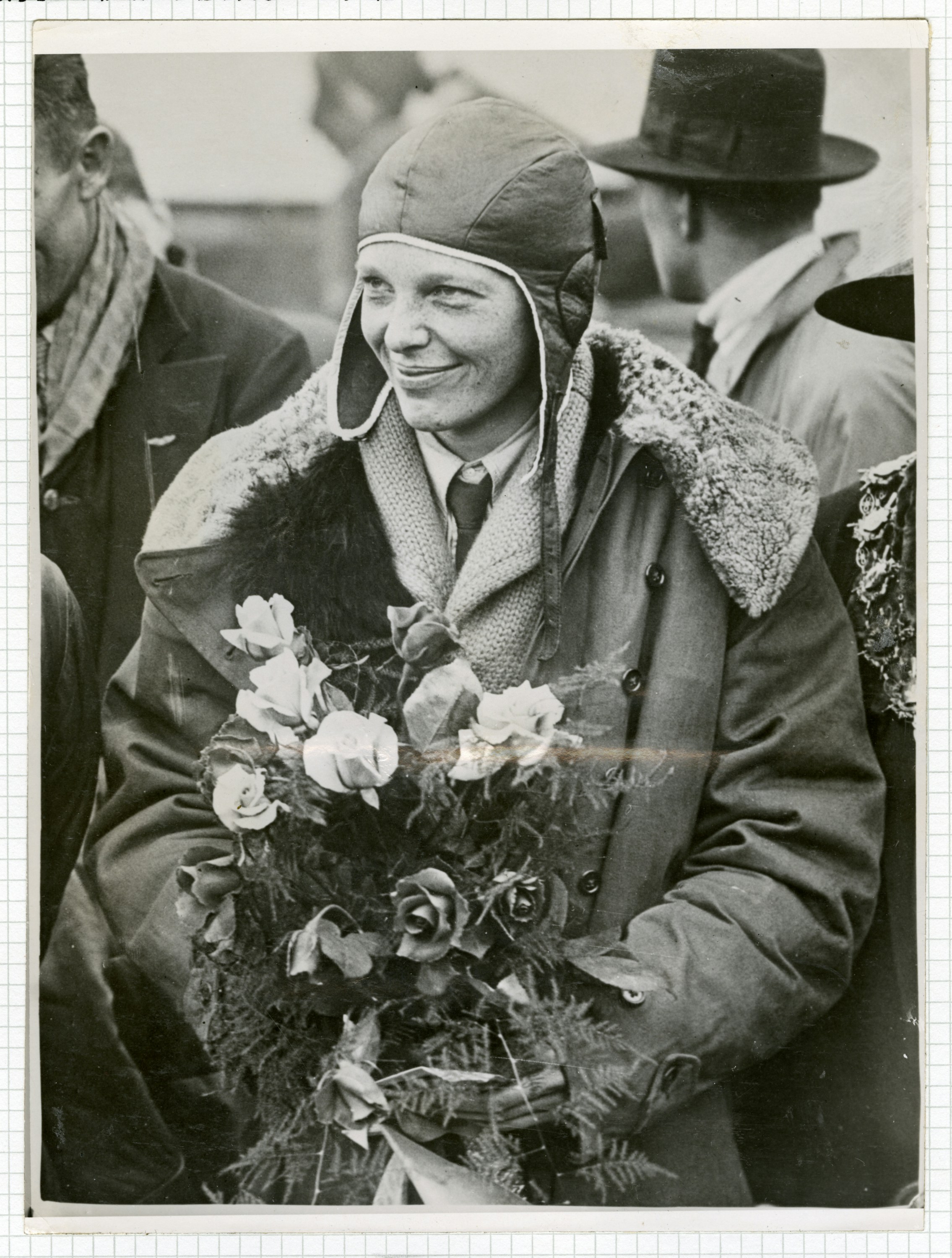 Woman in flight cap and coat smiles, holding flowers.