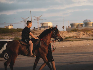 A boy rides a brown horse led by another person.