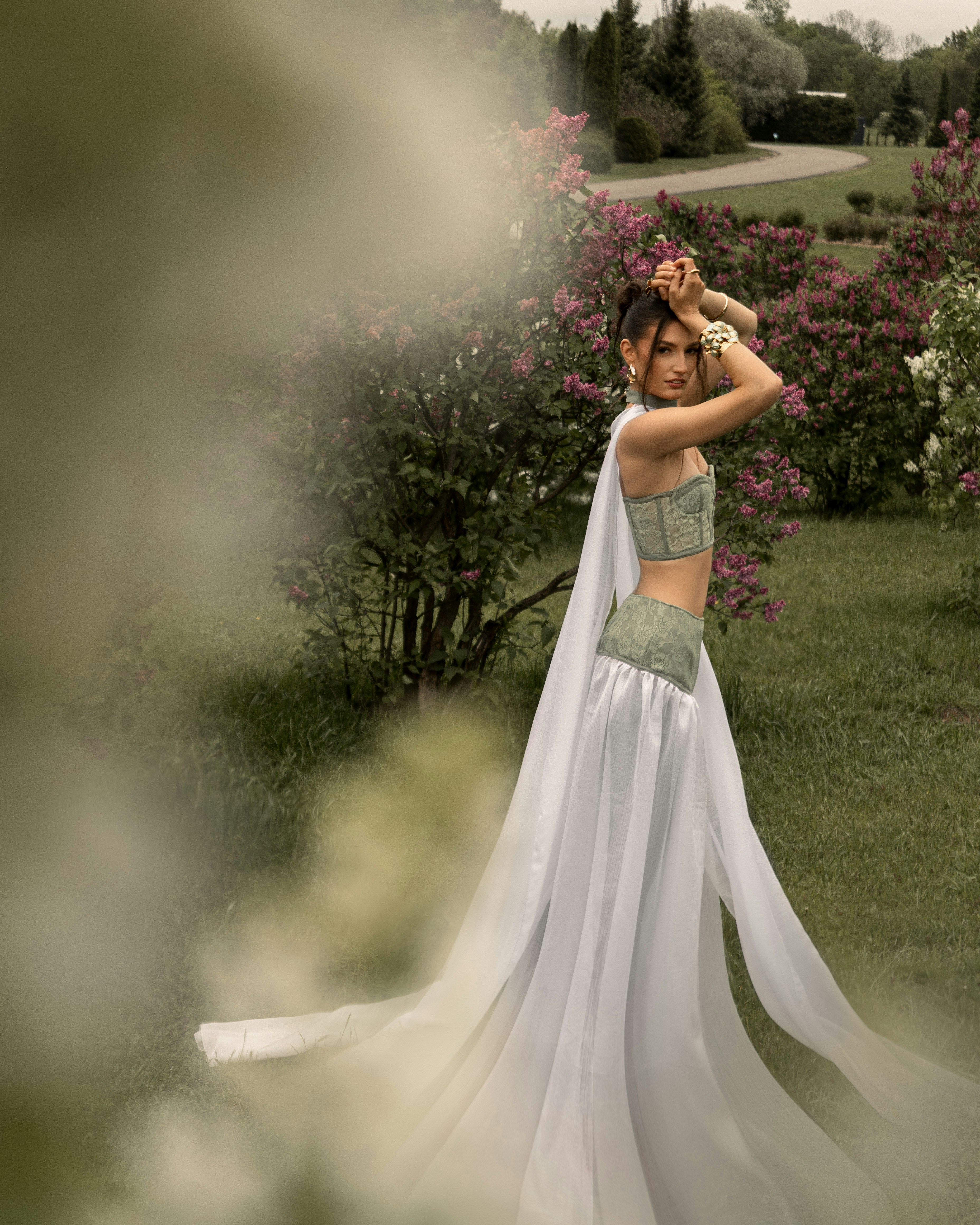 Mulher de vestido esvoaçante posa entre arbustos de lilás.