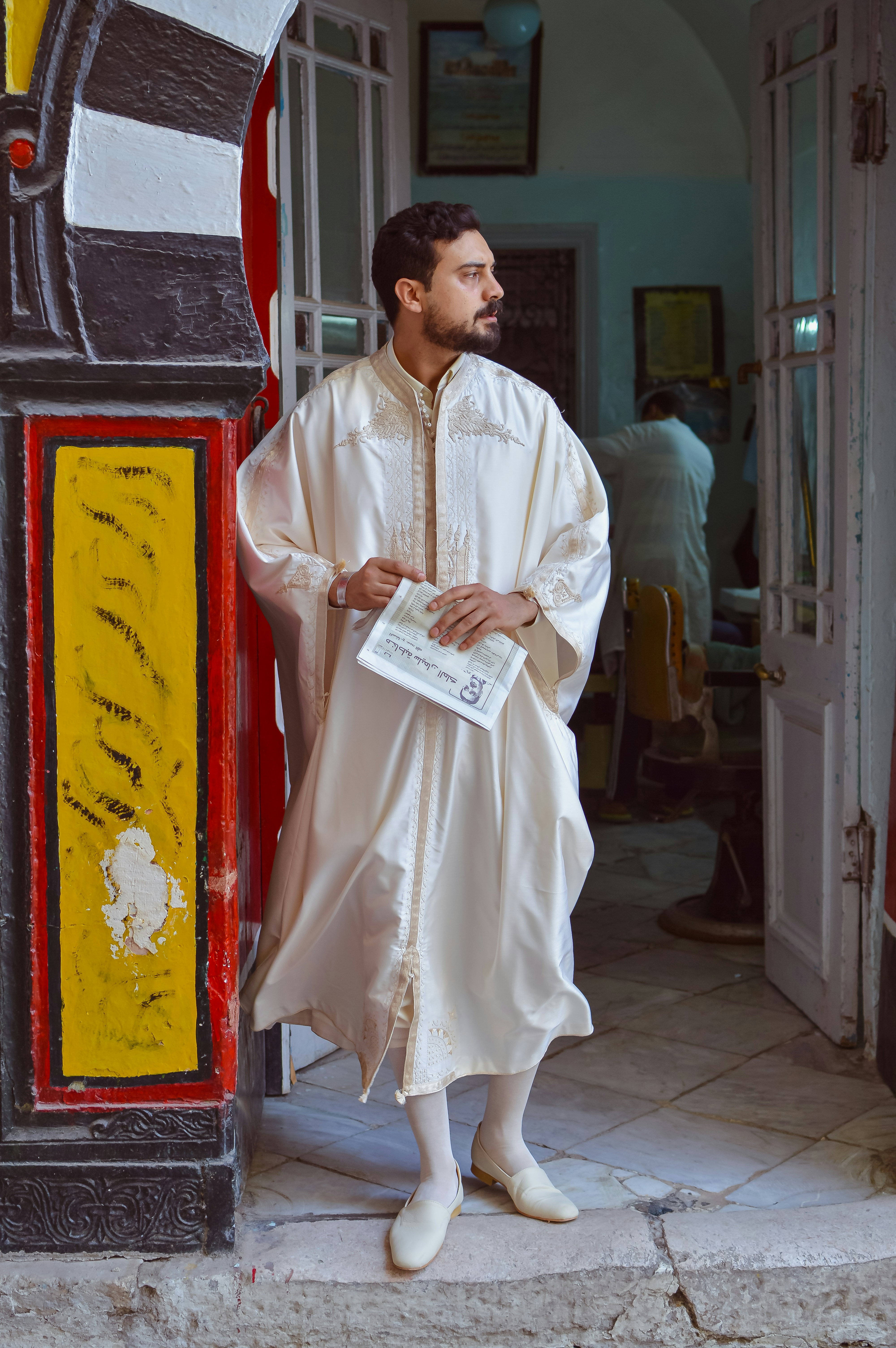 Man in traditional white robe holding newspaper in a doorway.