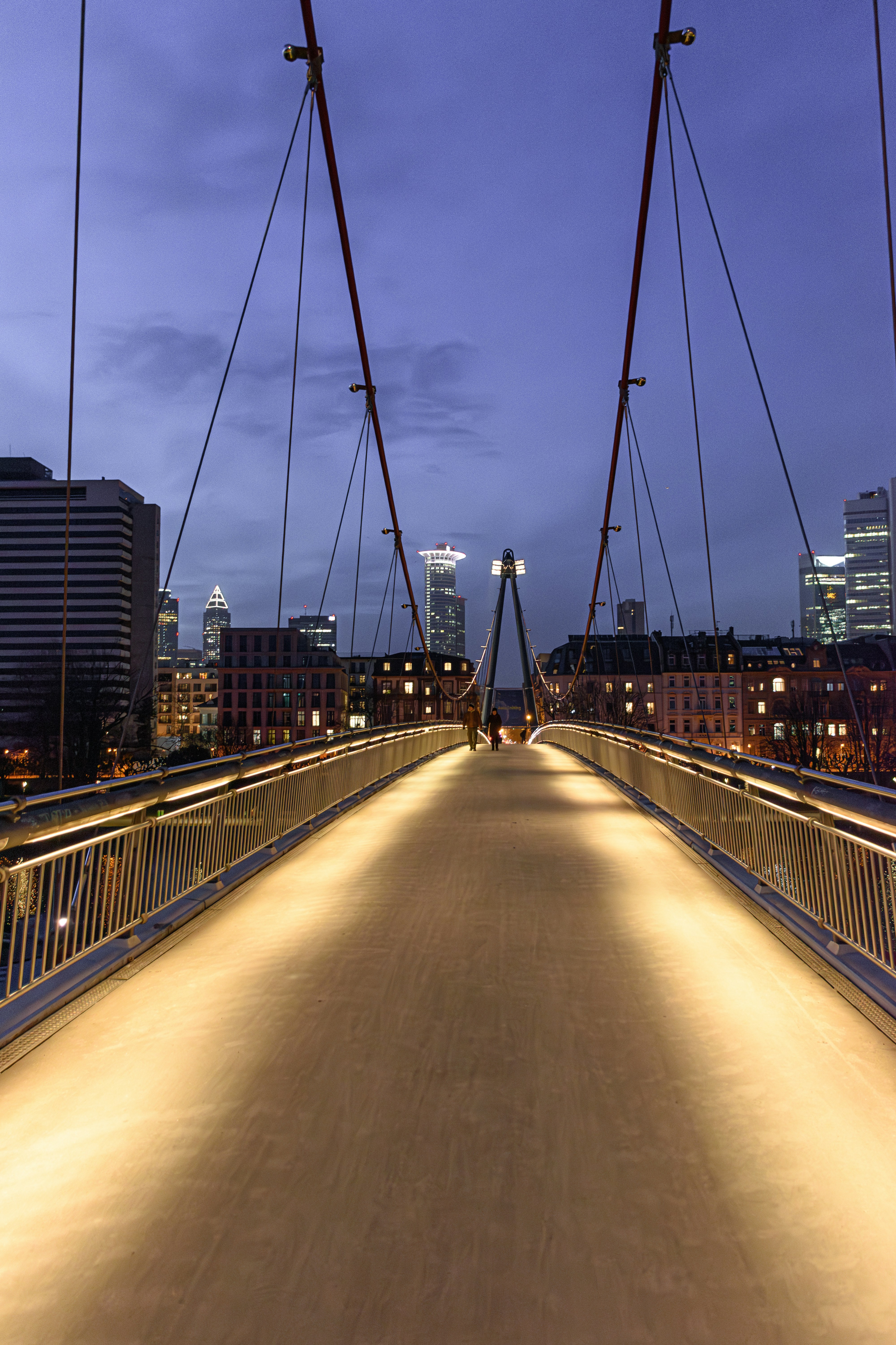 Illuminated pedestrian bridge leading to a city skyline at dusk.
