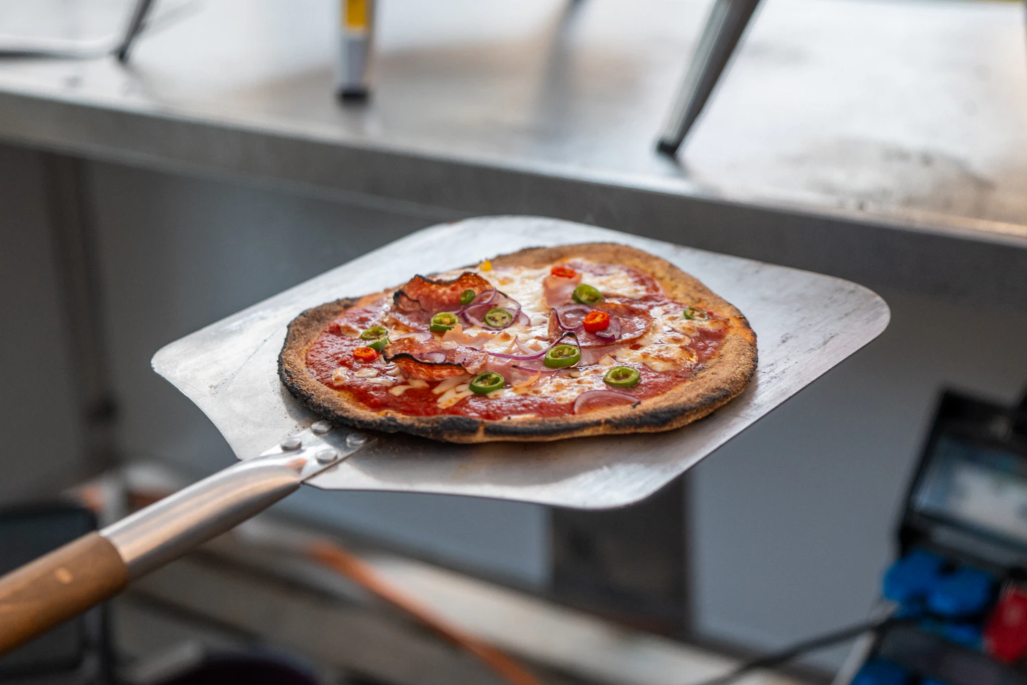 A small baked pizza held out on a perforated metal peel in a pizzeria kitchen