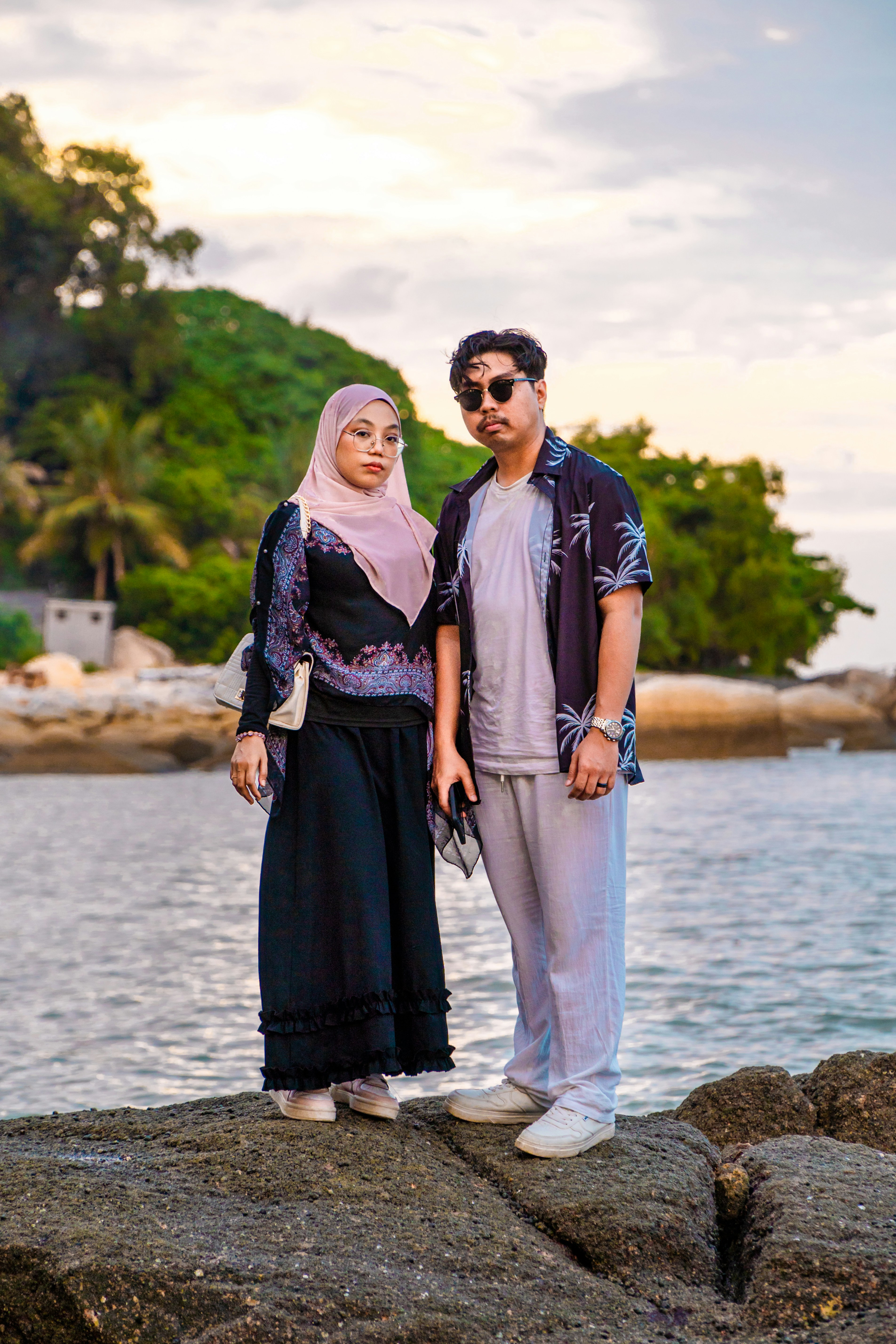 A couple standing on rocks by the sea at sunset.