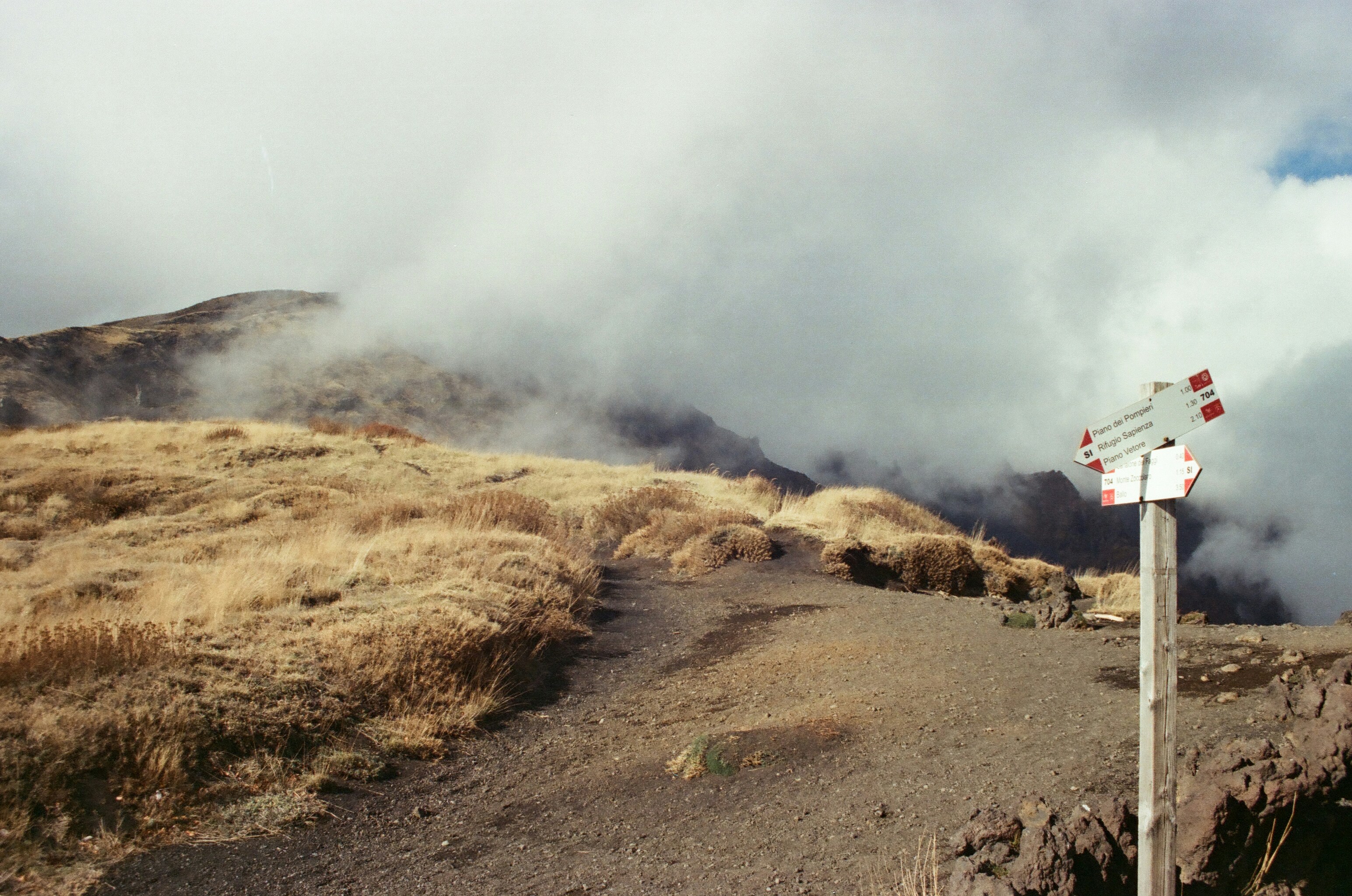 A mountain trail with dry grass, clouds, and a signpost