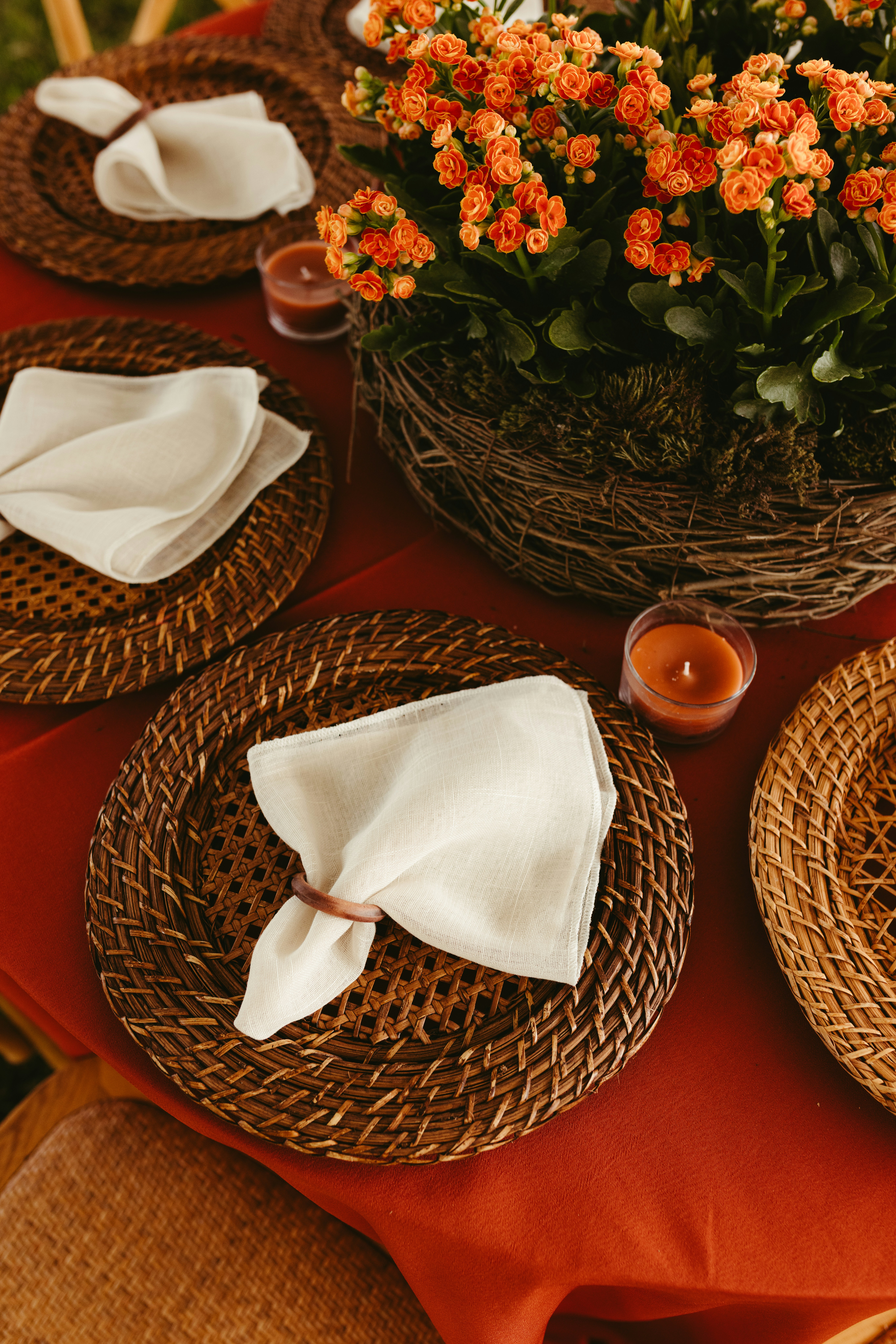 Tischdeckung mit gewebten Platzsets, weißen Servietten und orangefarbenen Blumen.