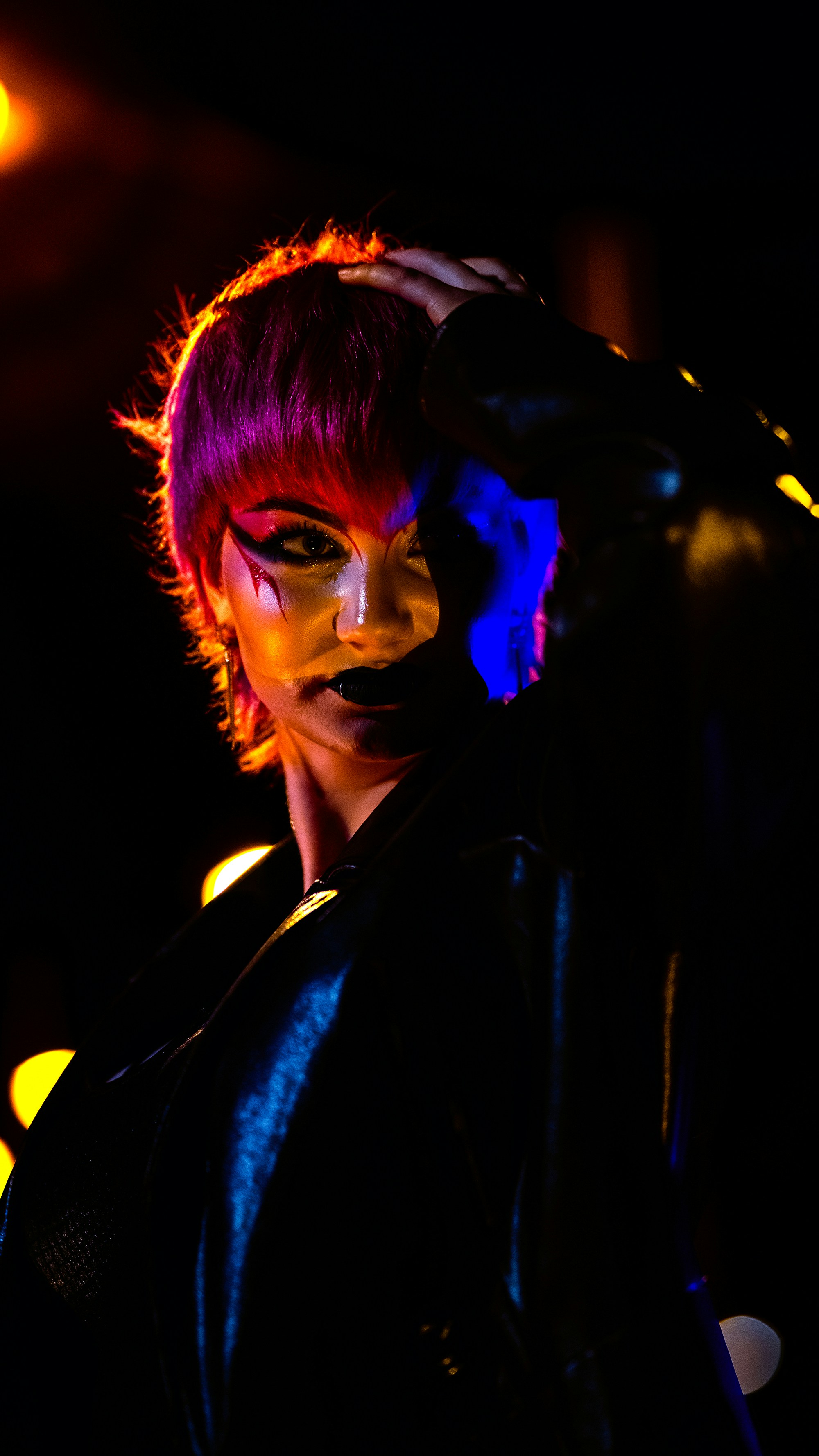 Orang dengan rambut magenta dan riasan dramatis di bawah lampu warna-warni.