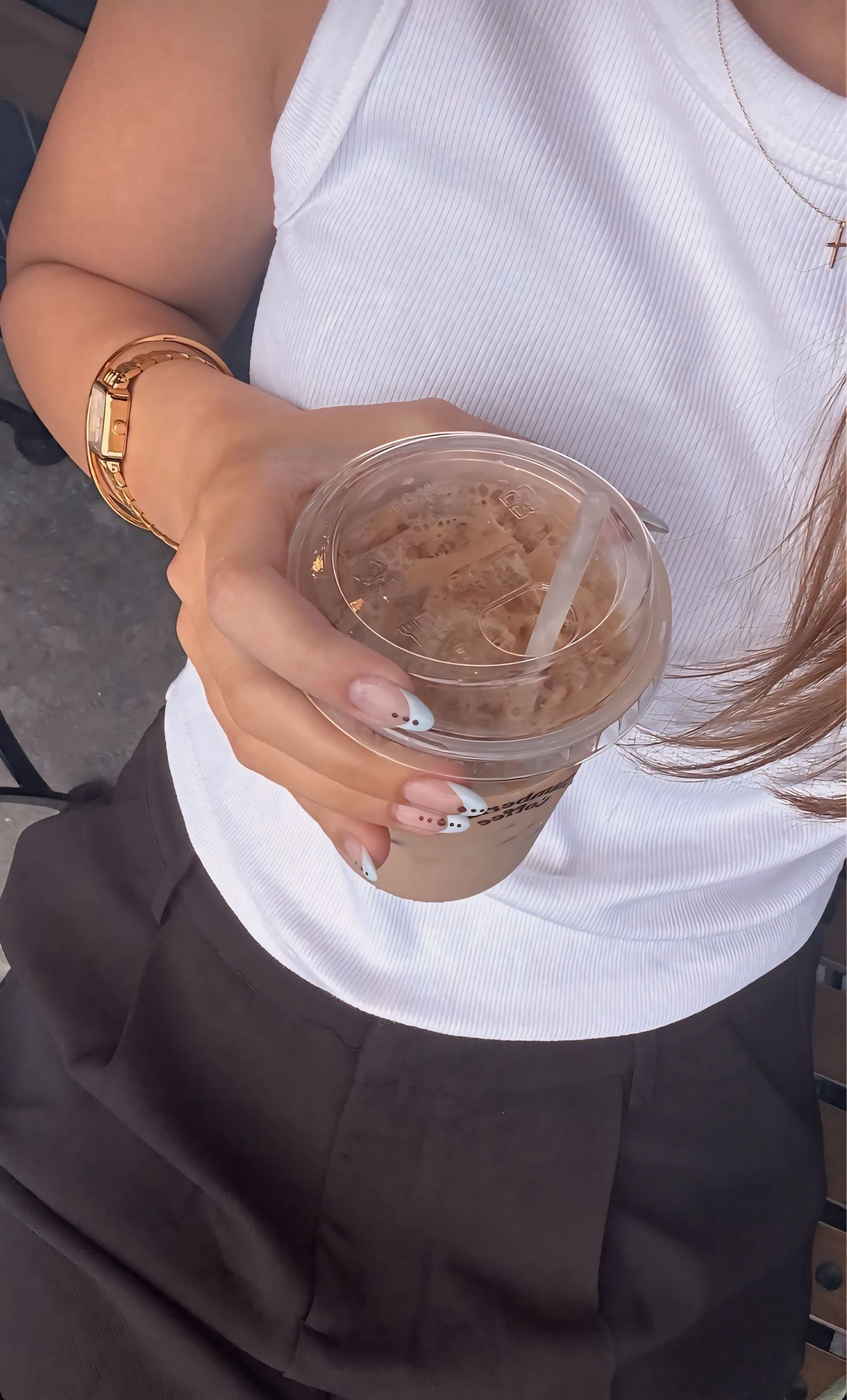 Overhead view of an iced coffee on a café table beside a woman in a white ribbed top wearing gold jewellery