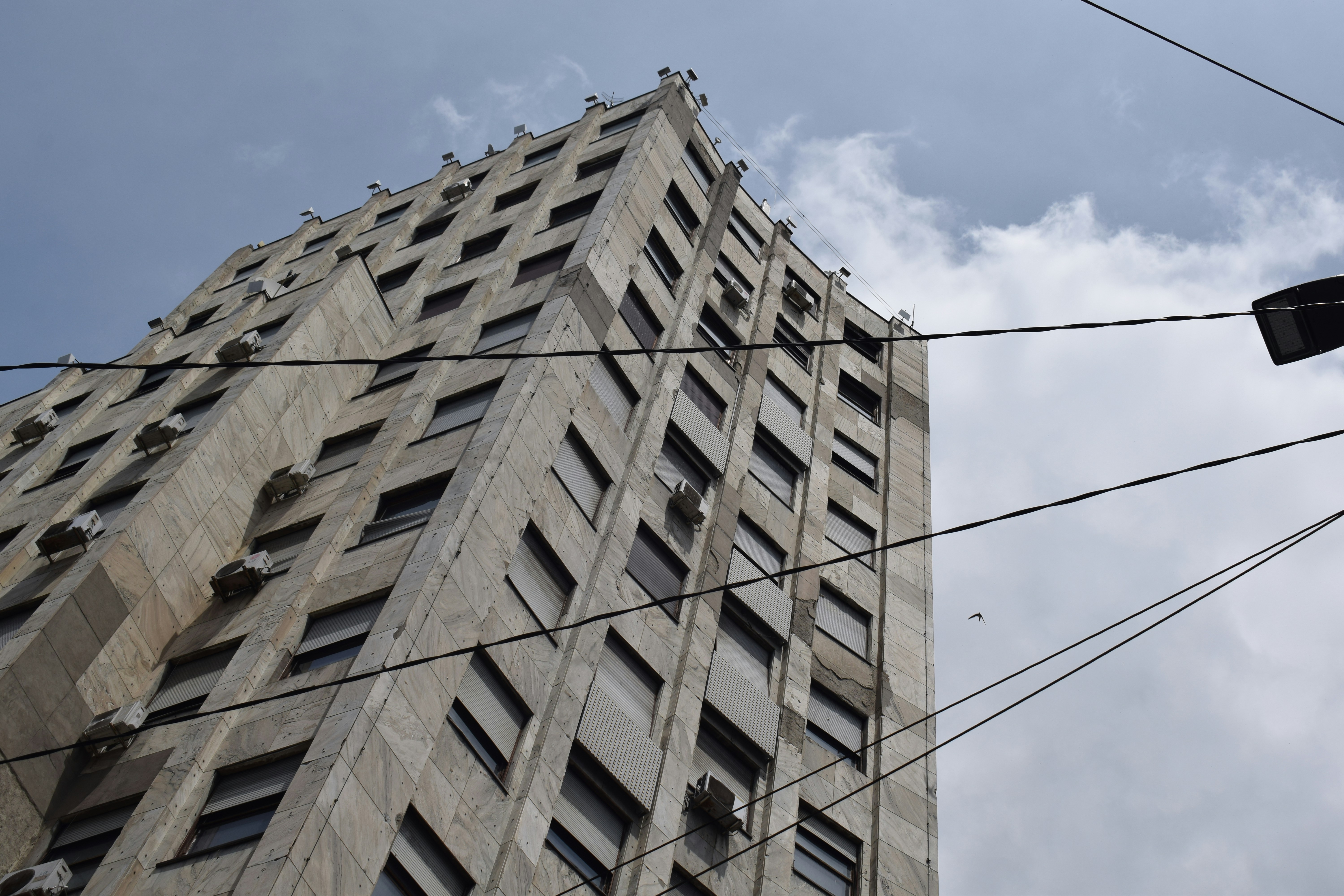 Prédio de cor clara com muitas janelas e fios sob céu nublado.