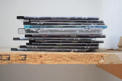 A stack of compact disc cases on a wooden shelf.