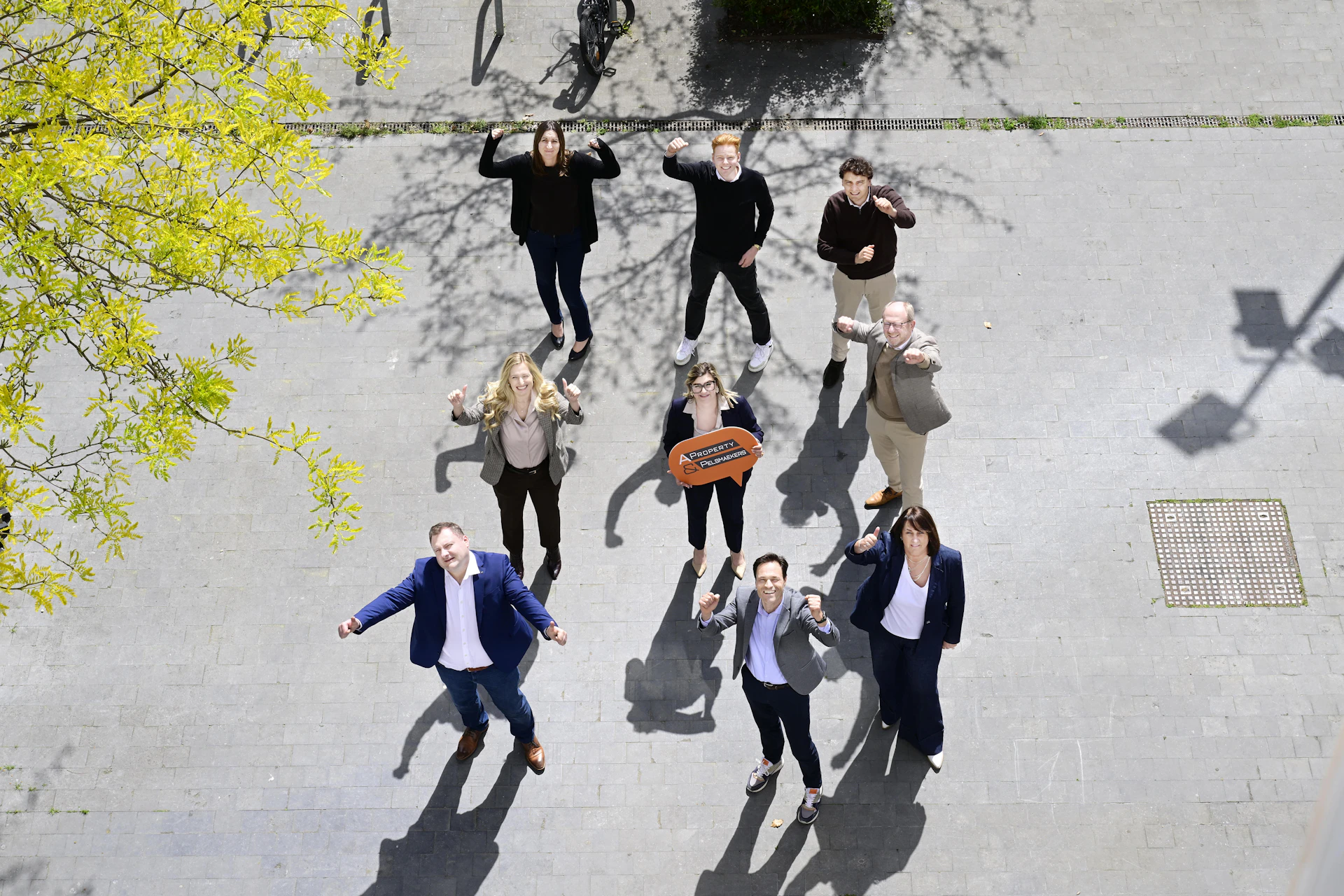 Nine people stand on pavement, looking up with raised arms