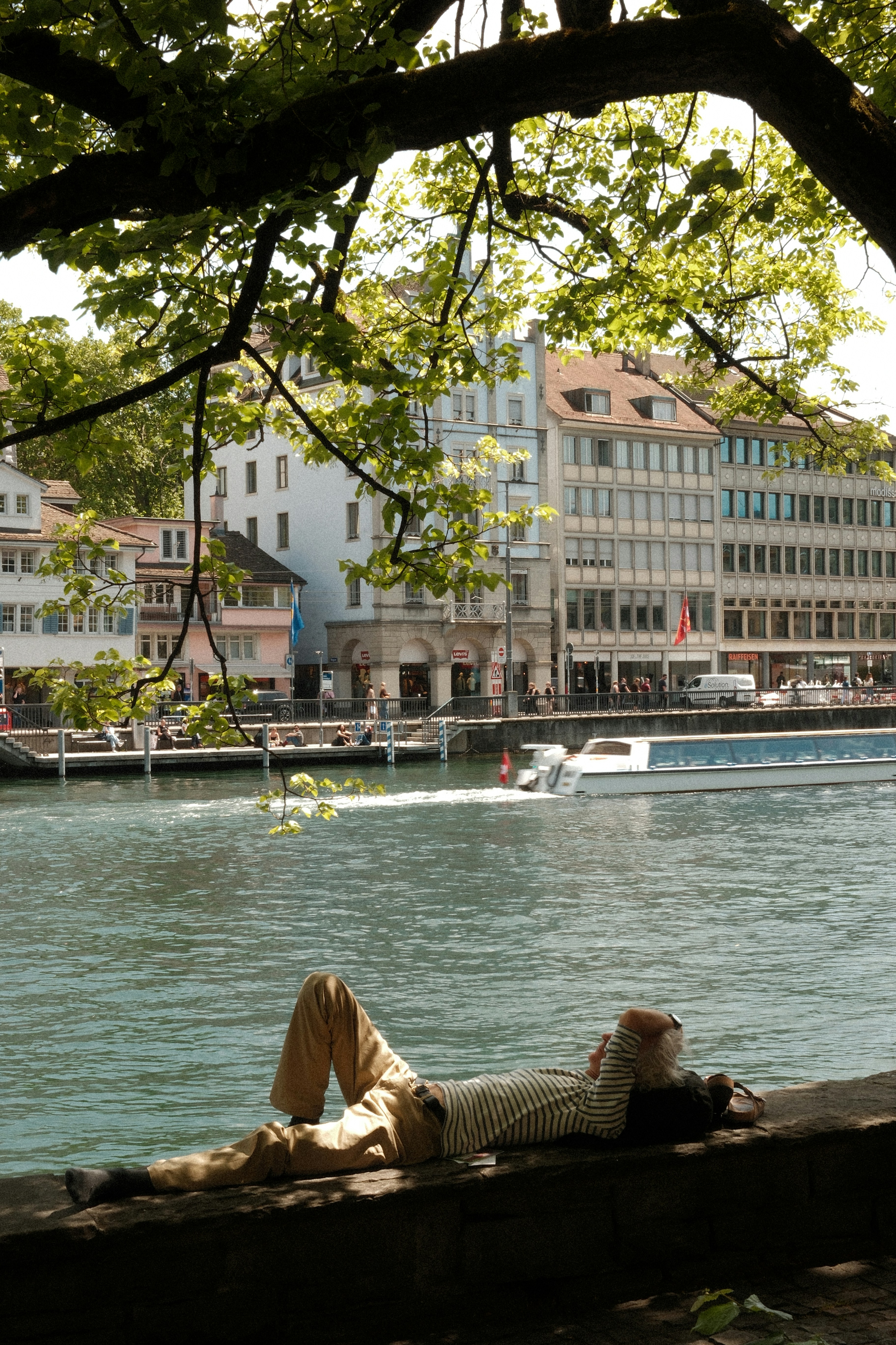 Person lying by river, city buildings and boat in background.