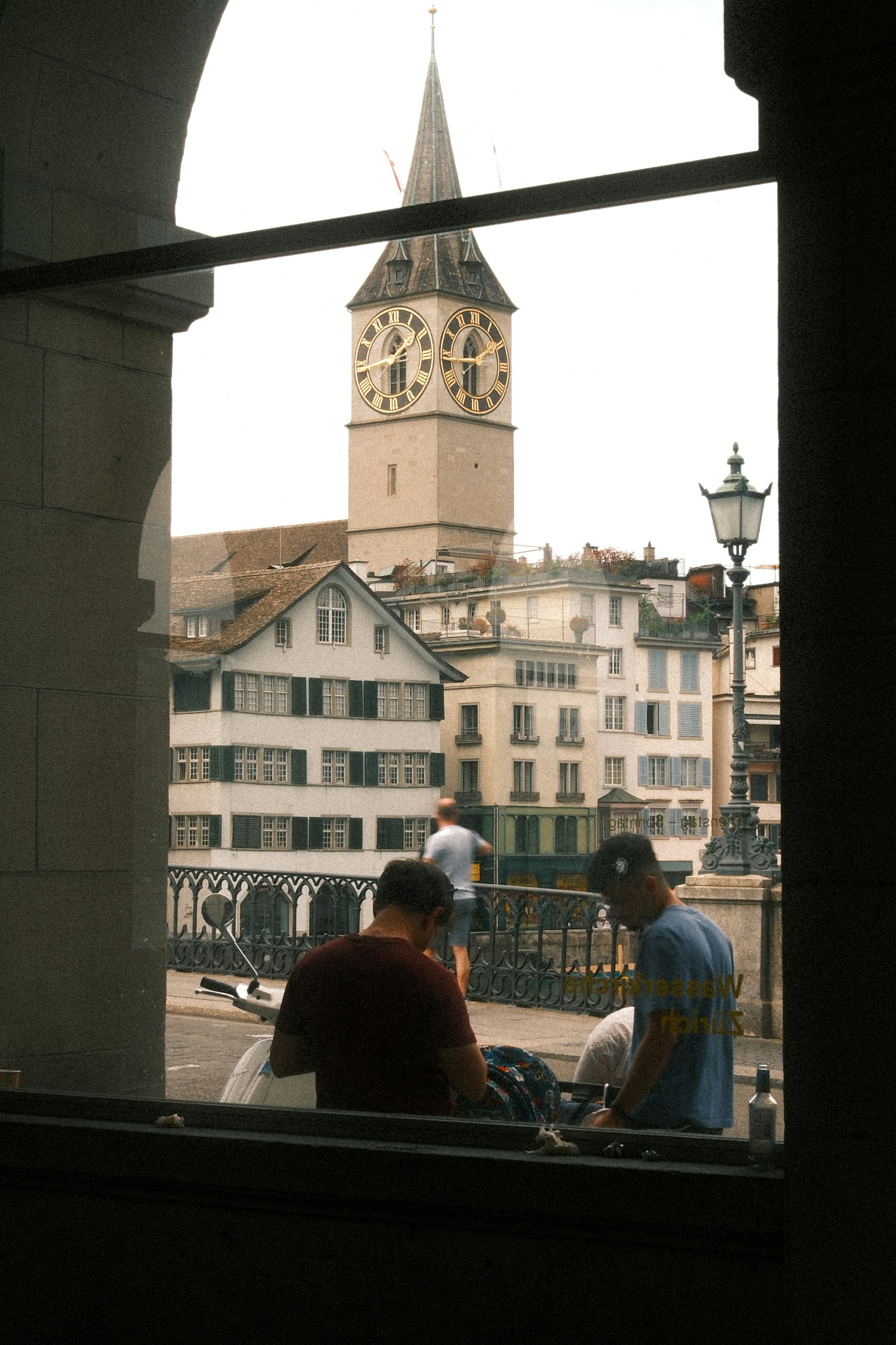 Two men sit overlooking a clock tower and city buildings.