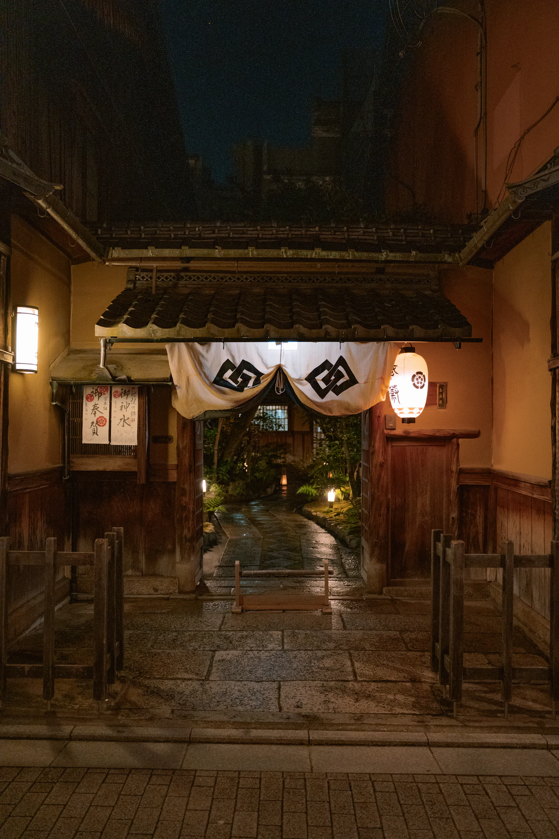 Traditional entrance with noren and glowing lanterns over a wet stone path at night