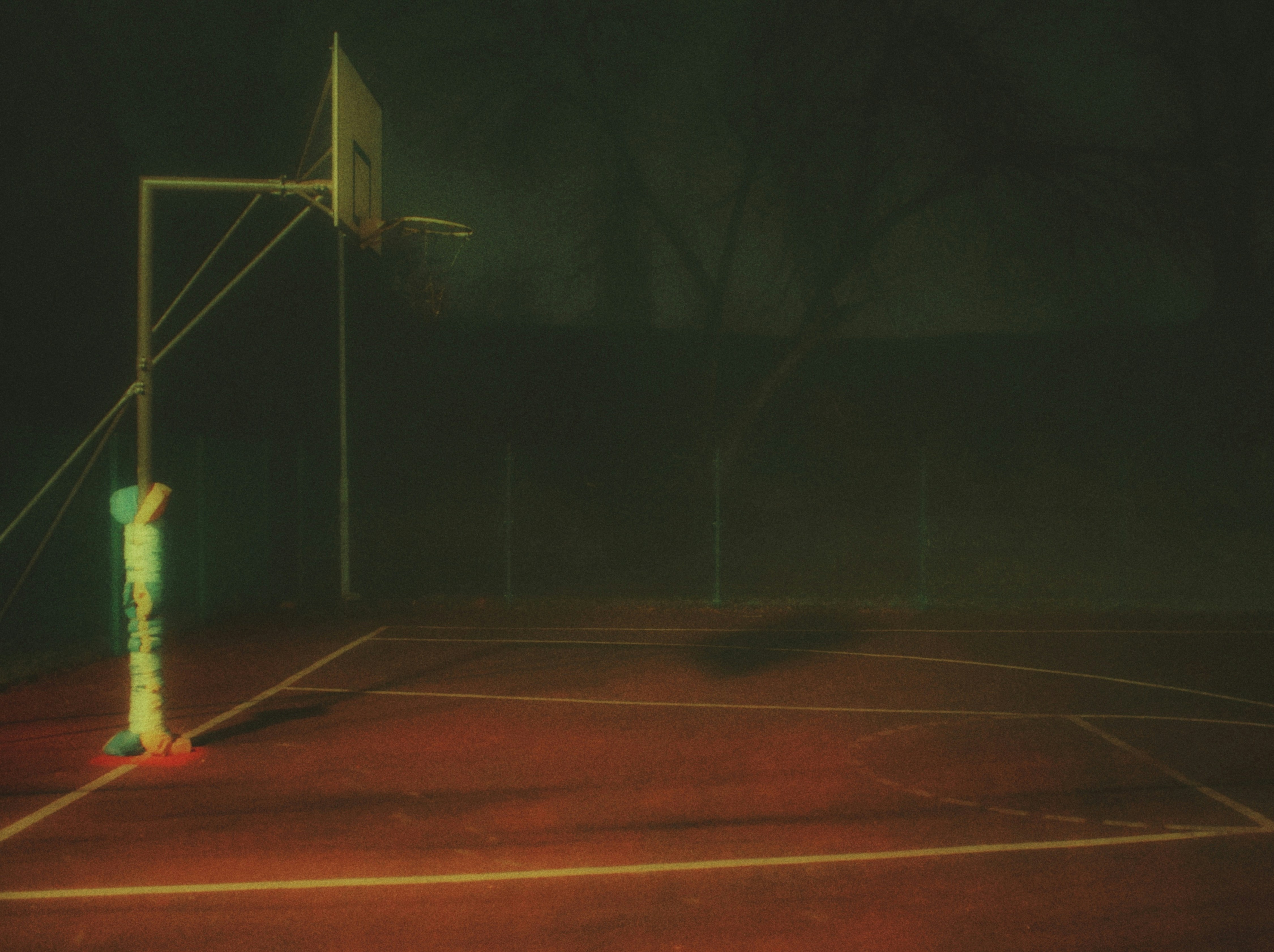 Foggy outdoor basketball court with a single hoop at night.