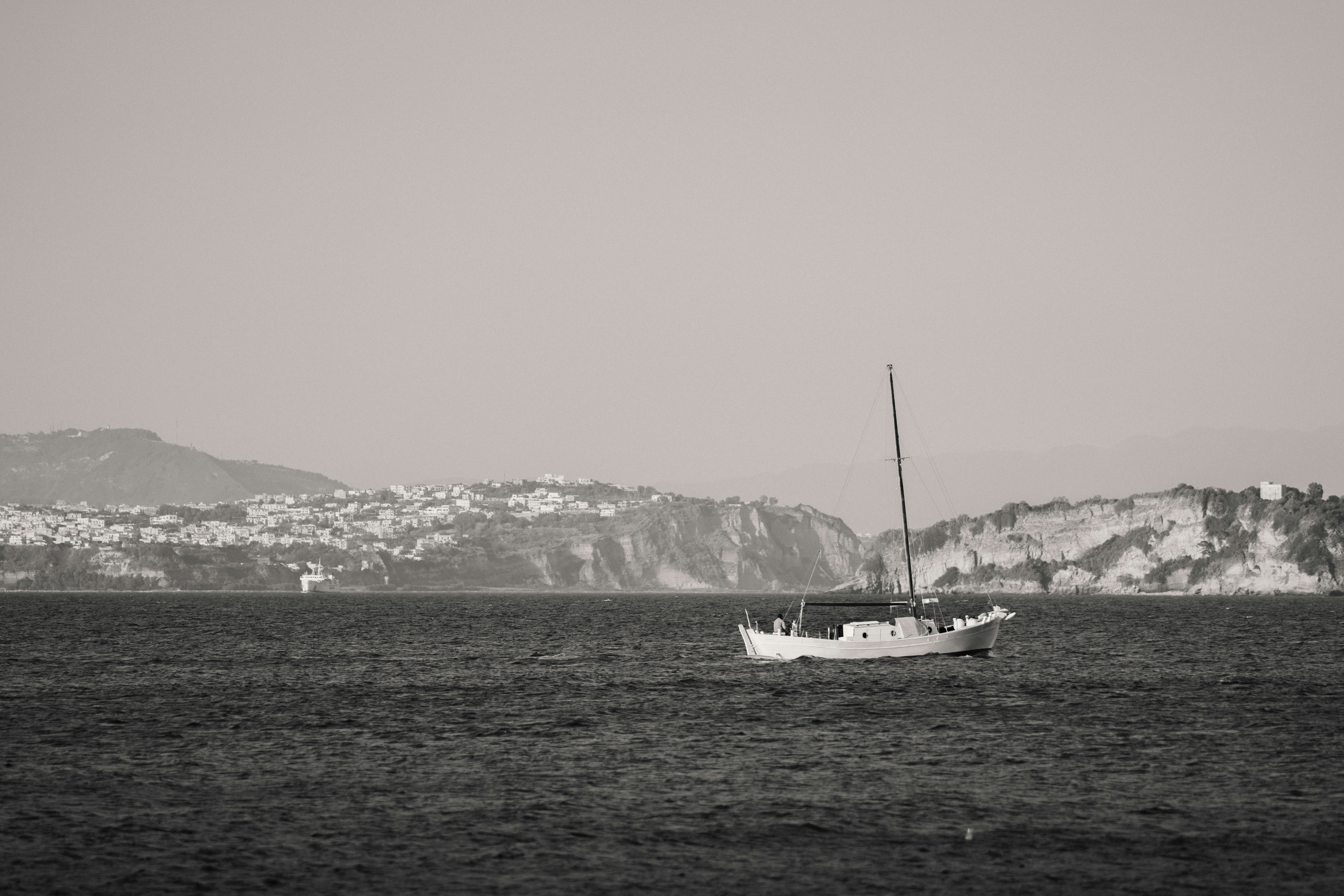 Monochrome sailboat on water with distant coastal town.