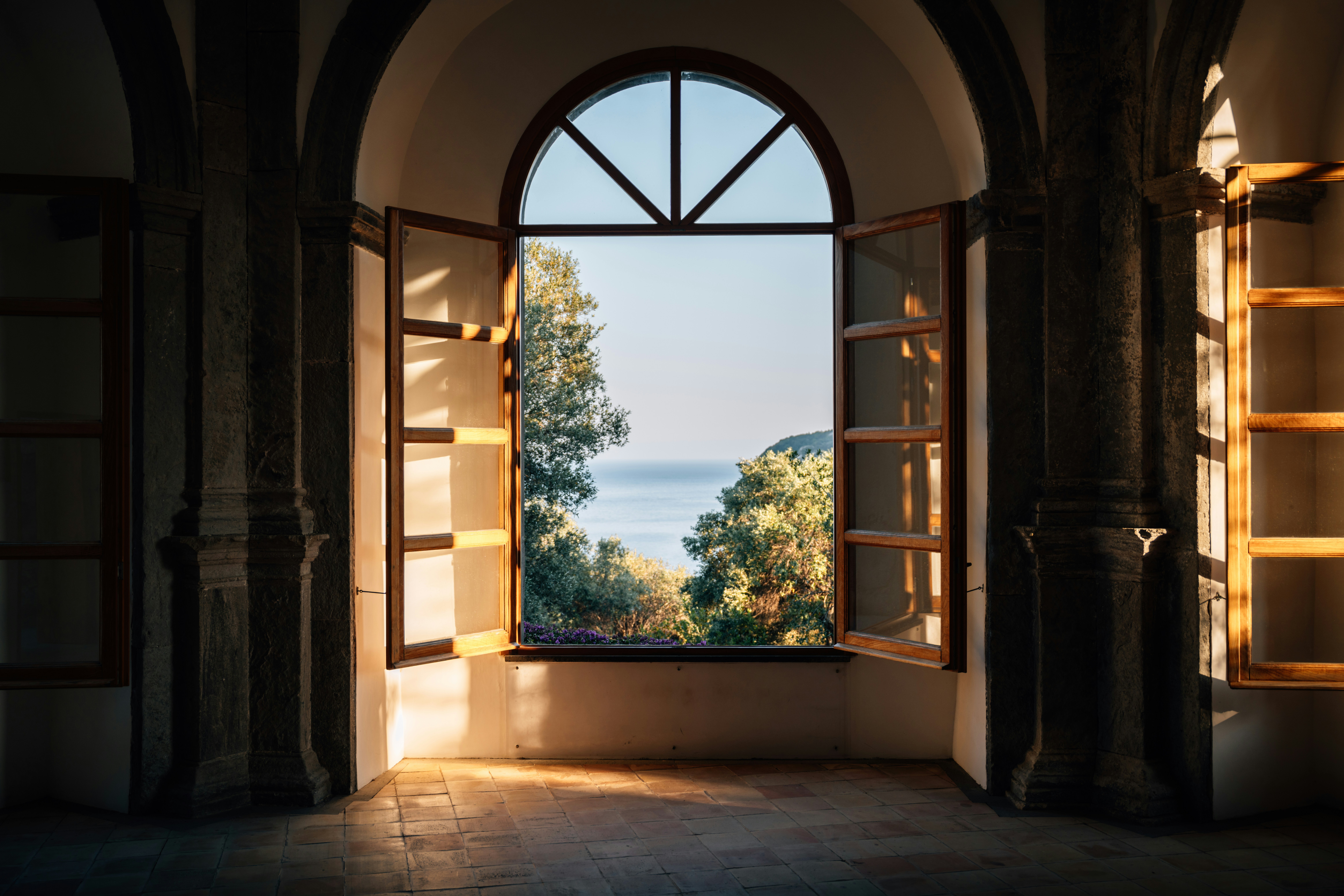 Sunlight streams through an open arched window overlooking the ocean.