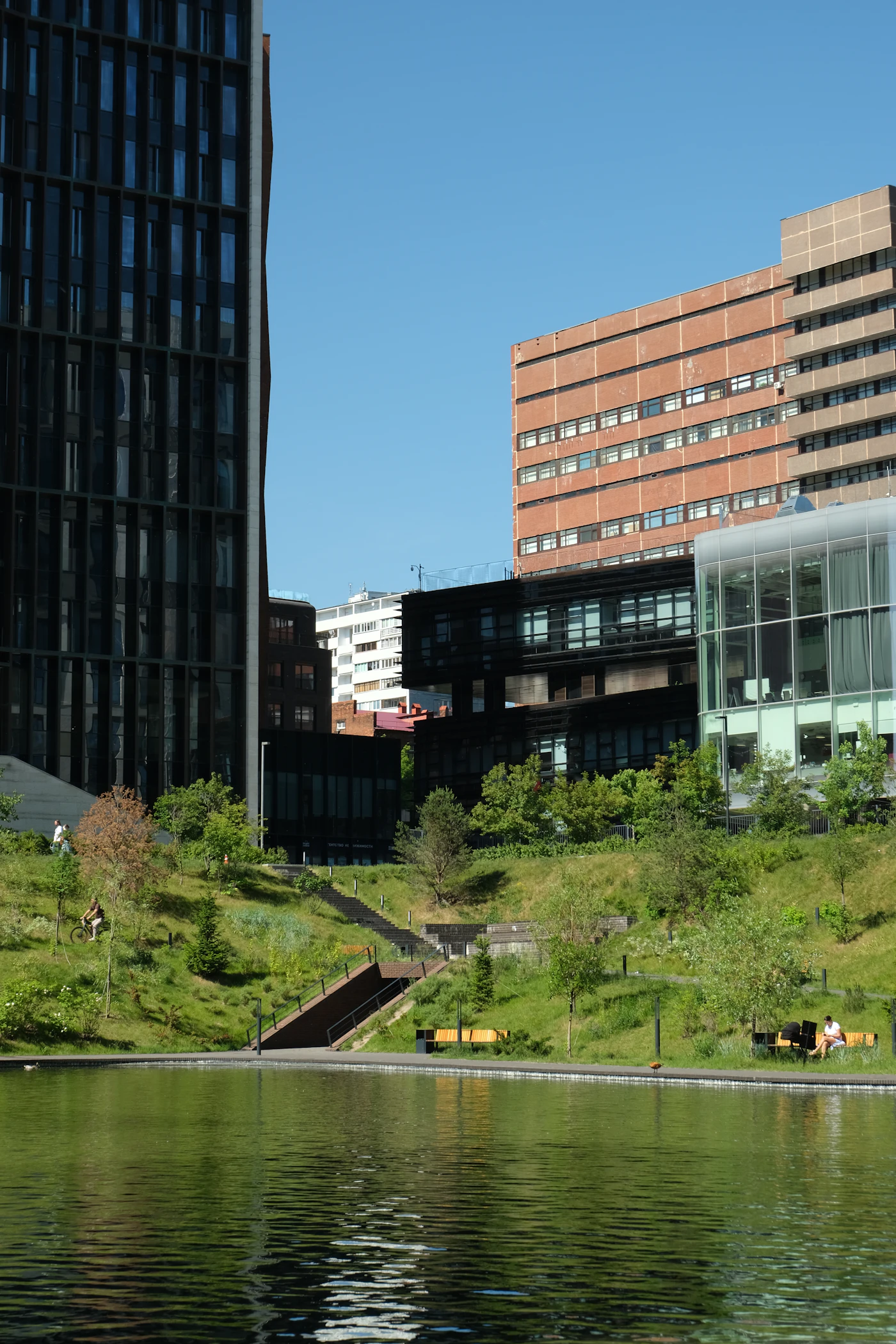People relaxing beside a pond and planted terraces surrounded by contemporary city buildings