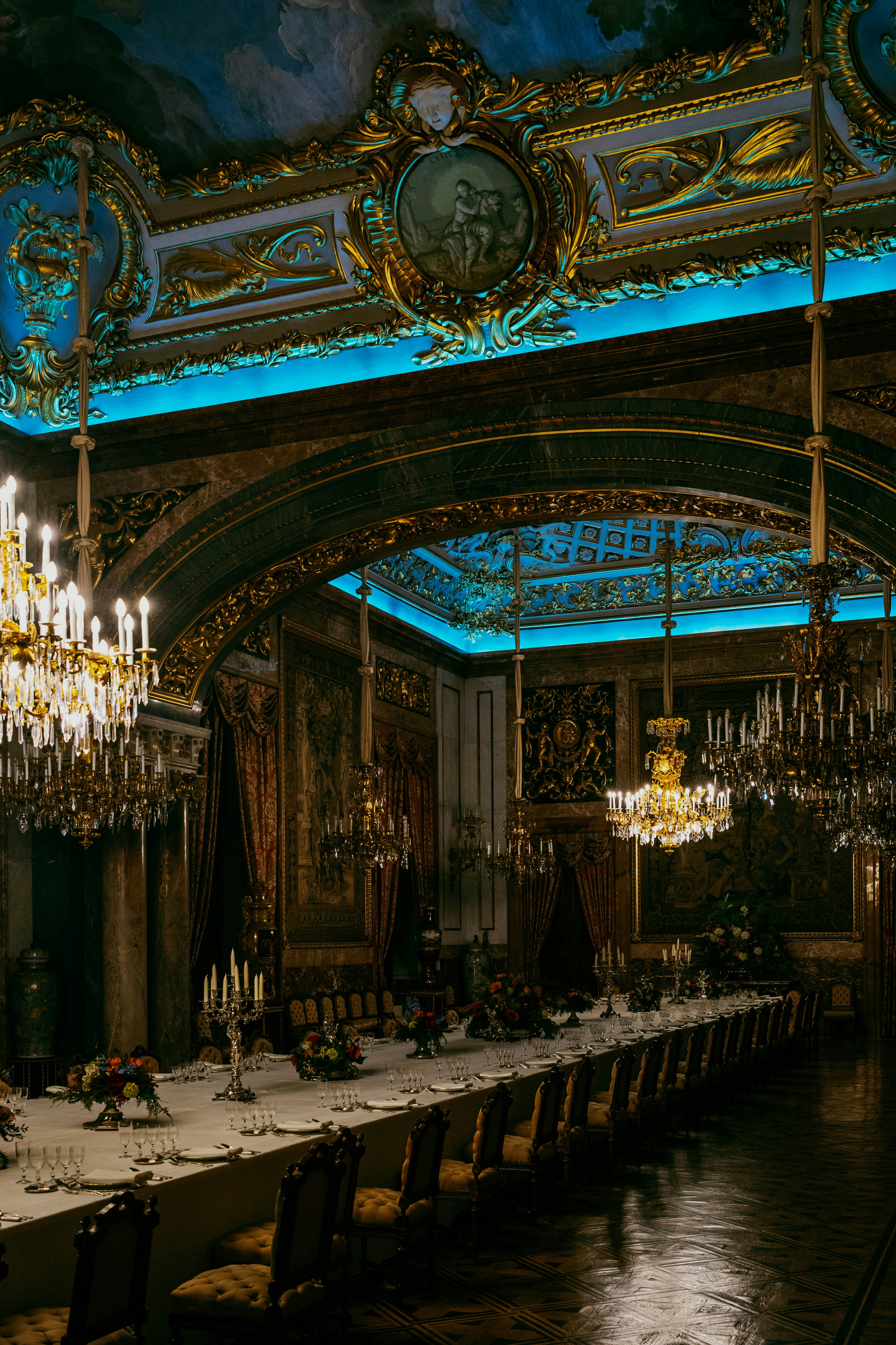 Opulent dining hall with chandeliers and a long, set table.