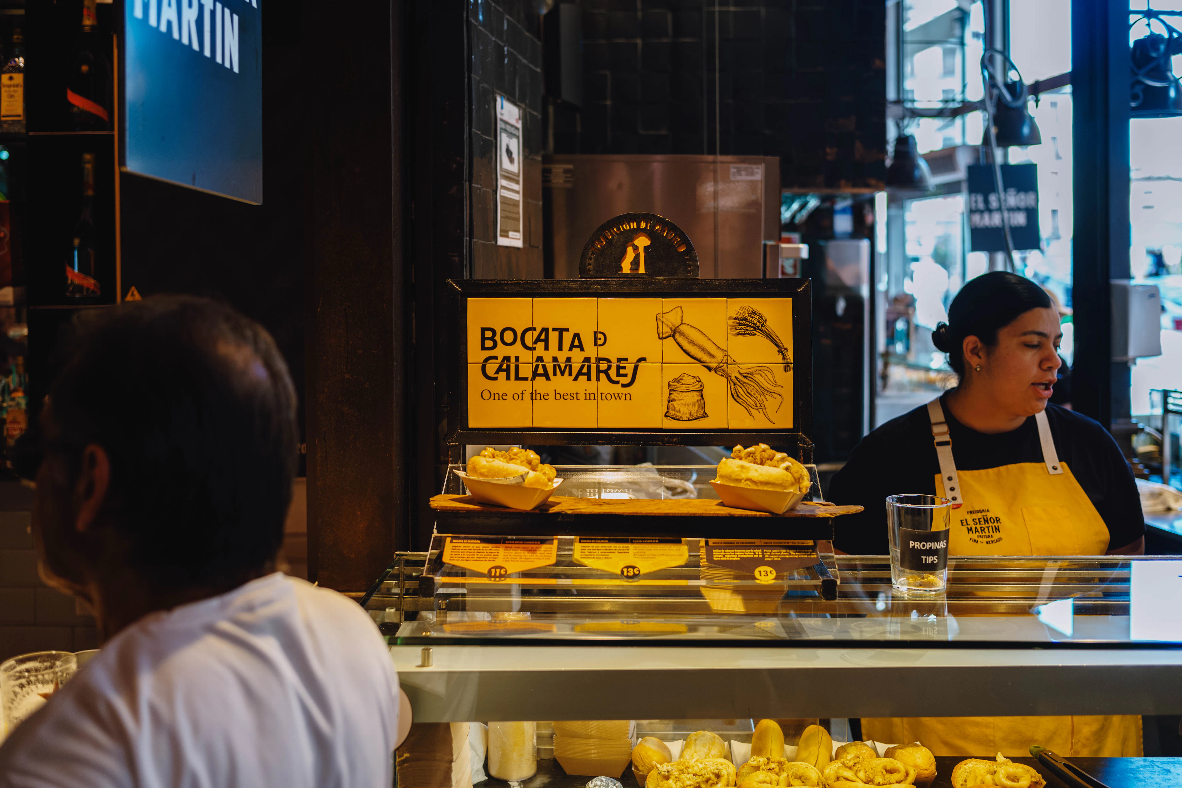 A vendor in a yellow apron behind a Madrid counter serving bocadillos de calamares, two fried-squid rolls waiting in paper trays under a hand-lettered Bocata de Calamares sign.