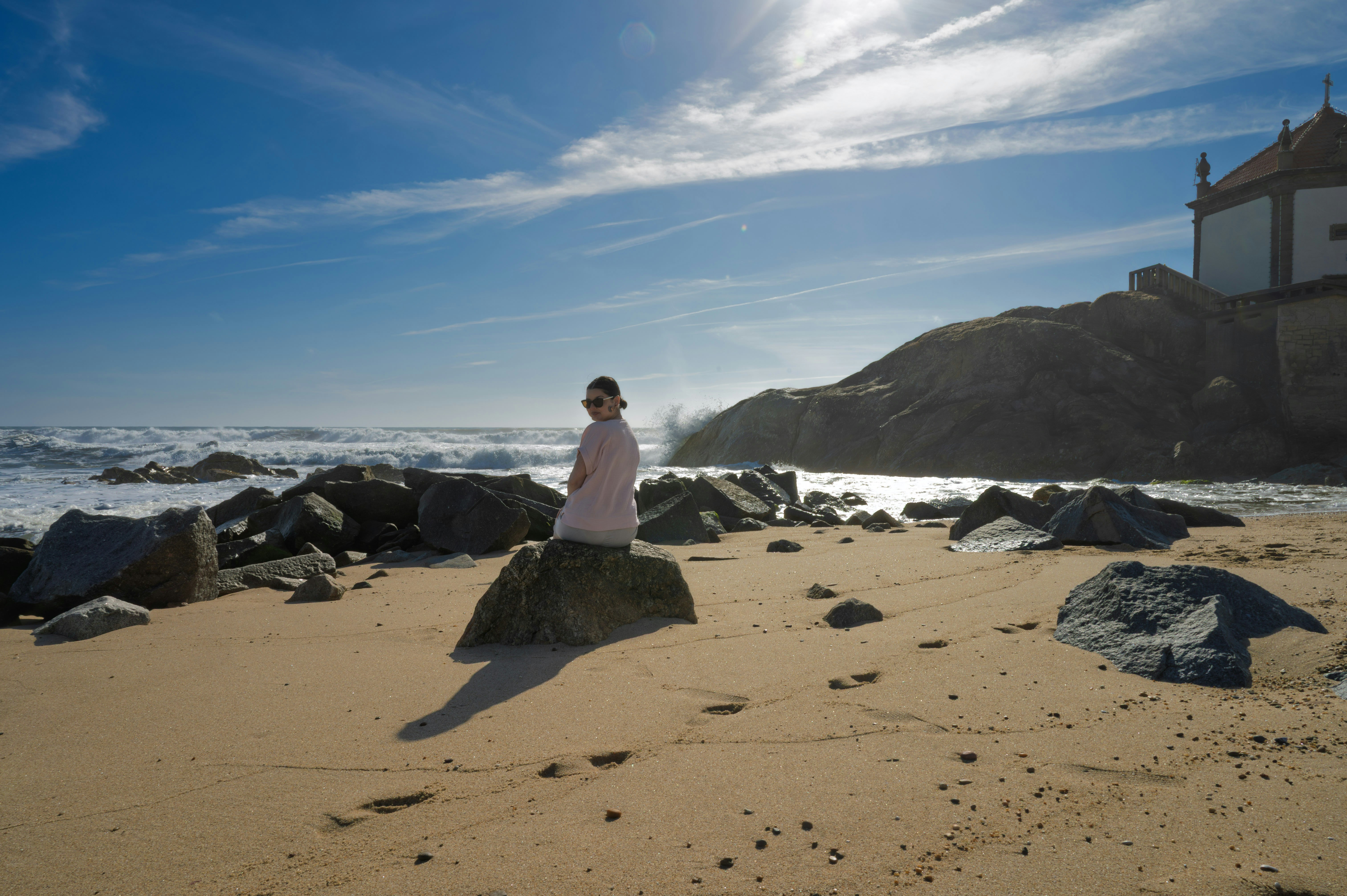 Person on a sunny, rocky beach with ocean and building.