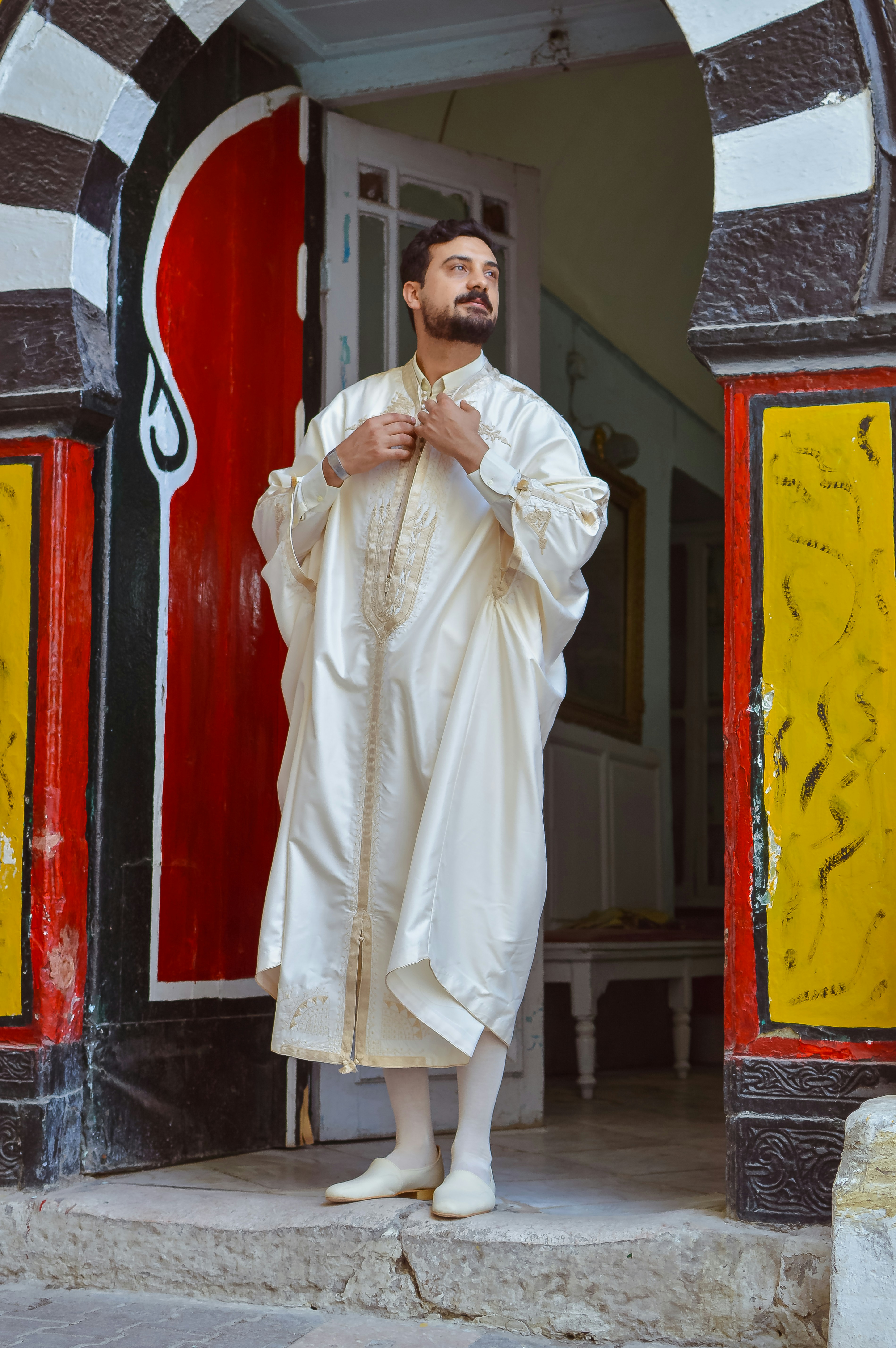 Man in elegant cream traditional outfit poses in ornate archway.