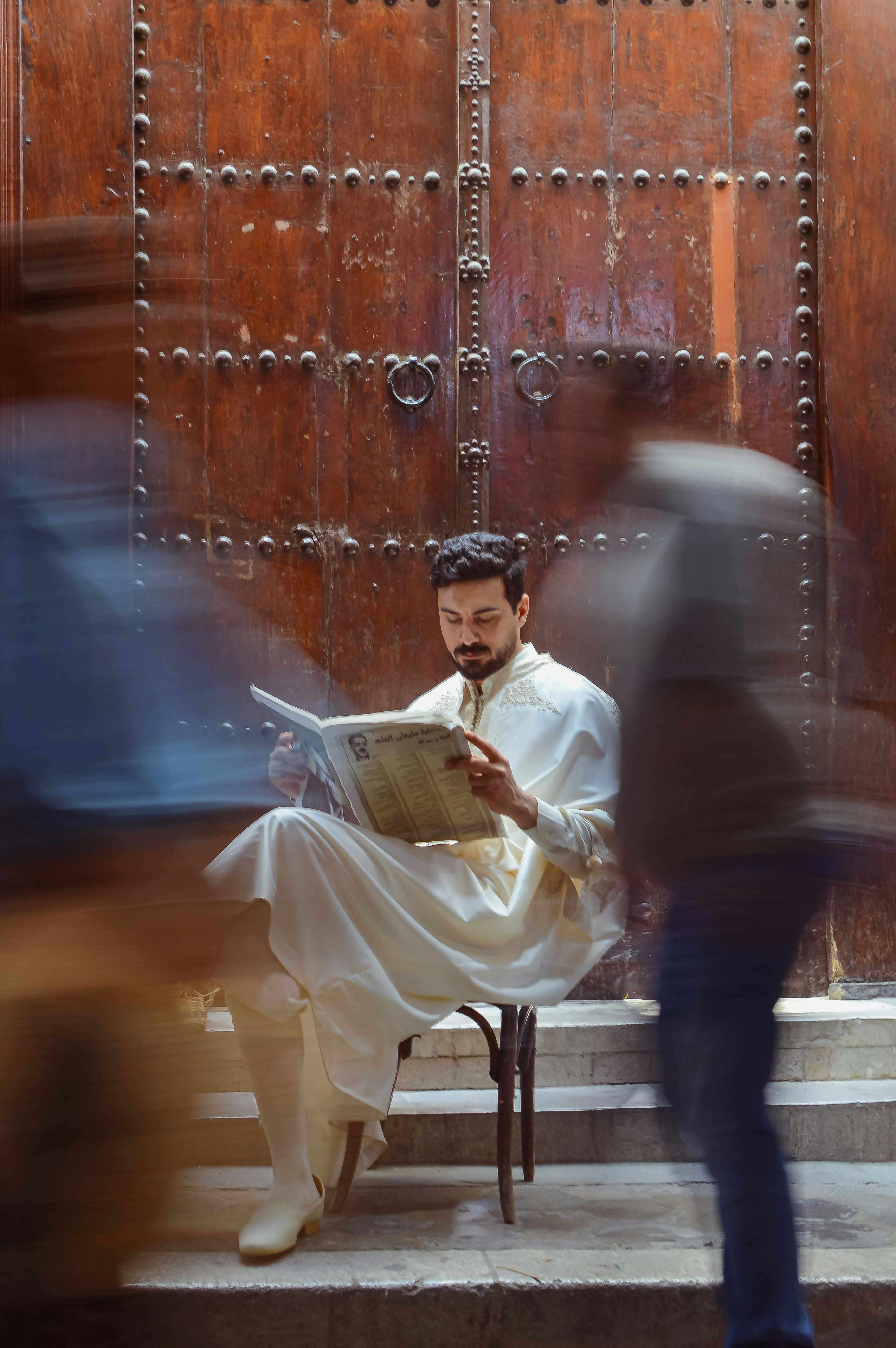 Man in traditional attire reads newspaper by ornate wooden door.