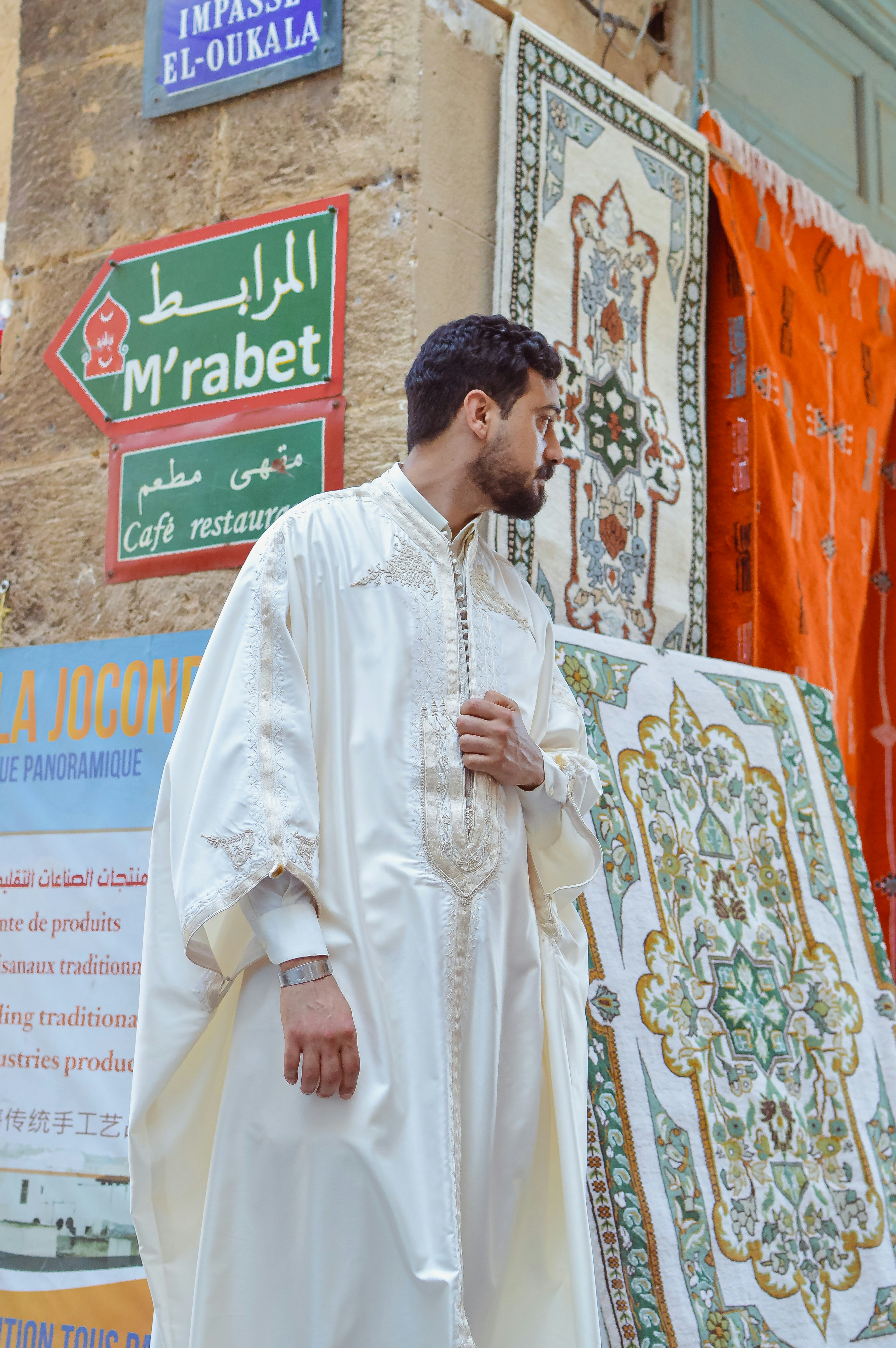Man in white traditional attire stands by colorful hanging rugs.