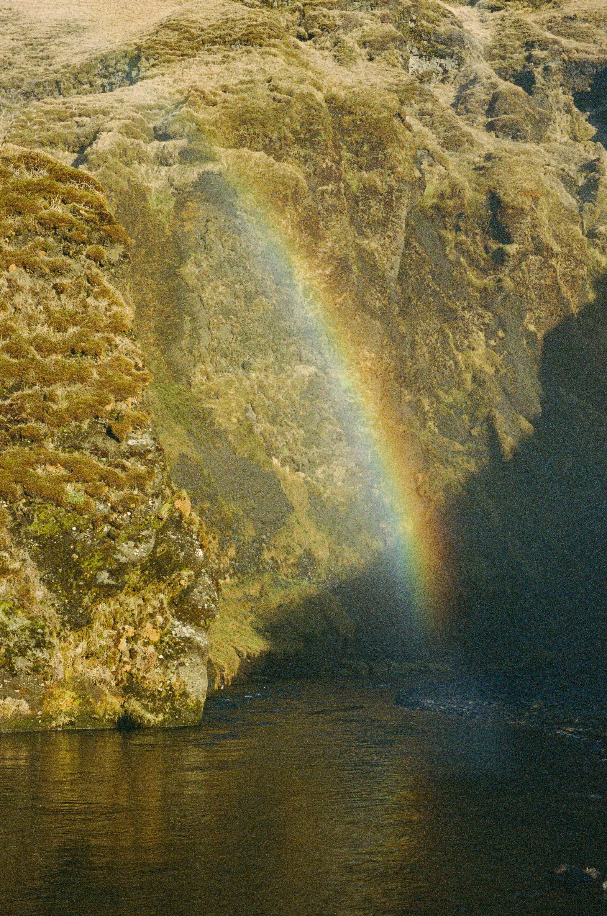Rainbow arches across a rugged cliff above a dark river.