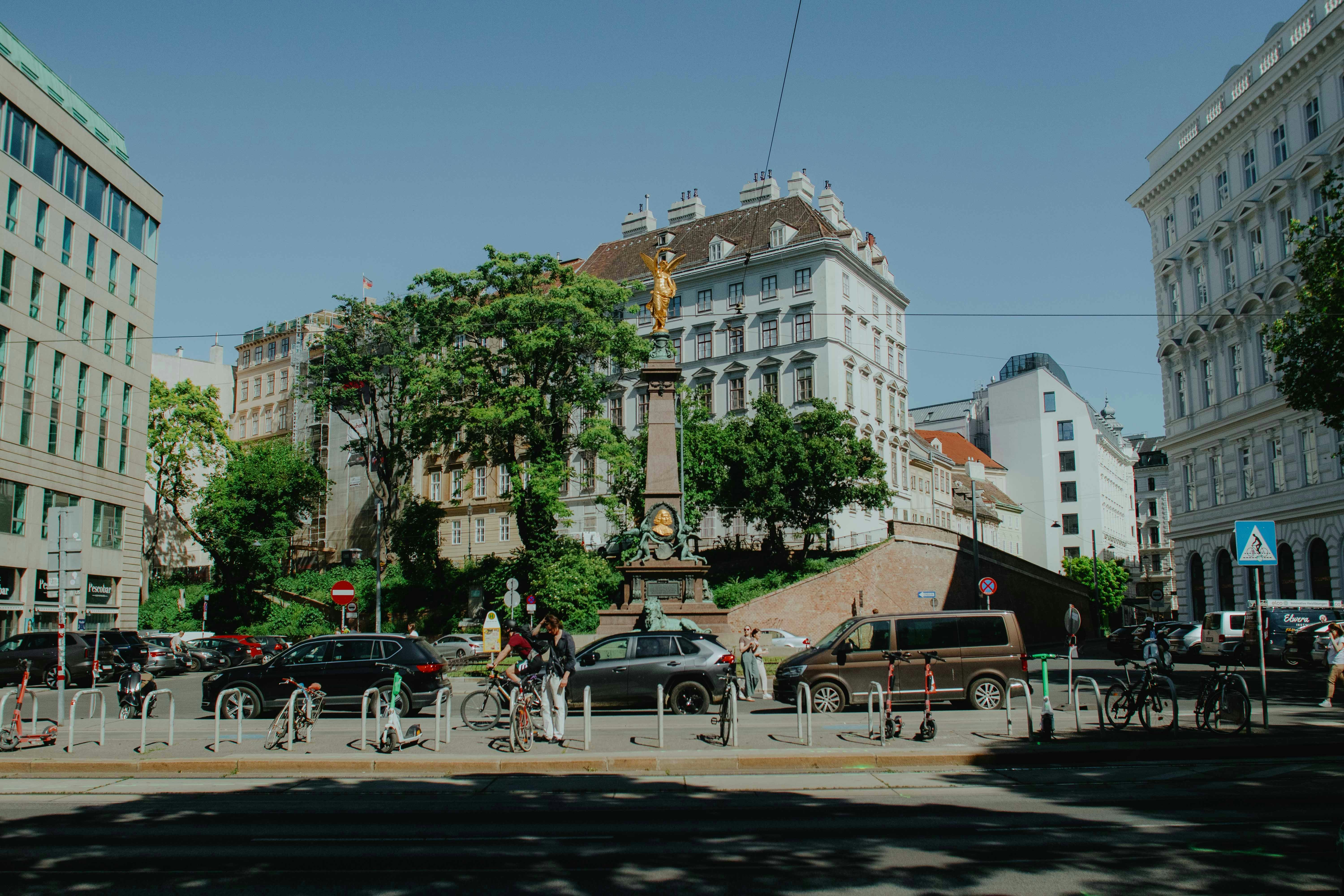 City square with monument, historic buildings, cars, and trees.