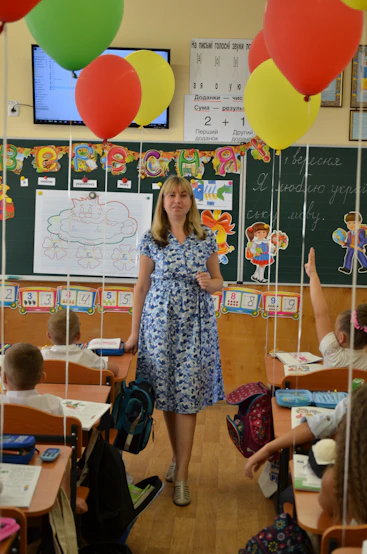 Teacher standing in a classroom with students and colorful balloons.