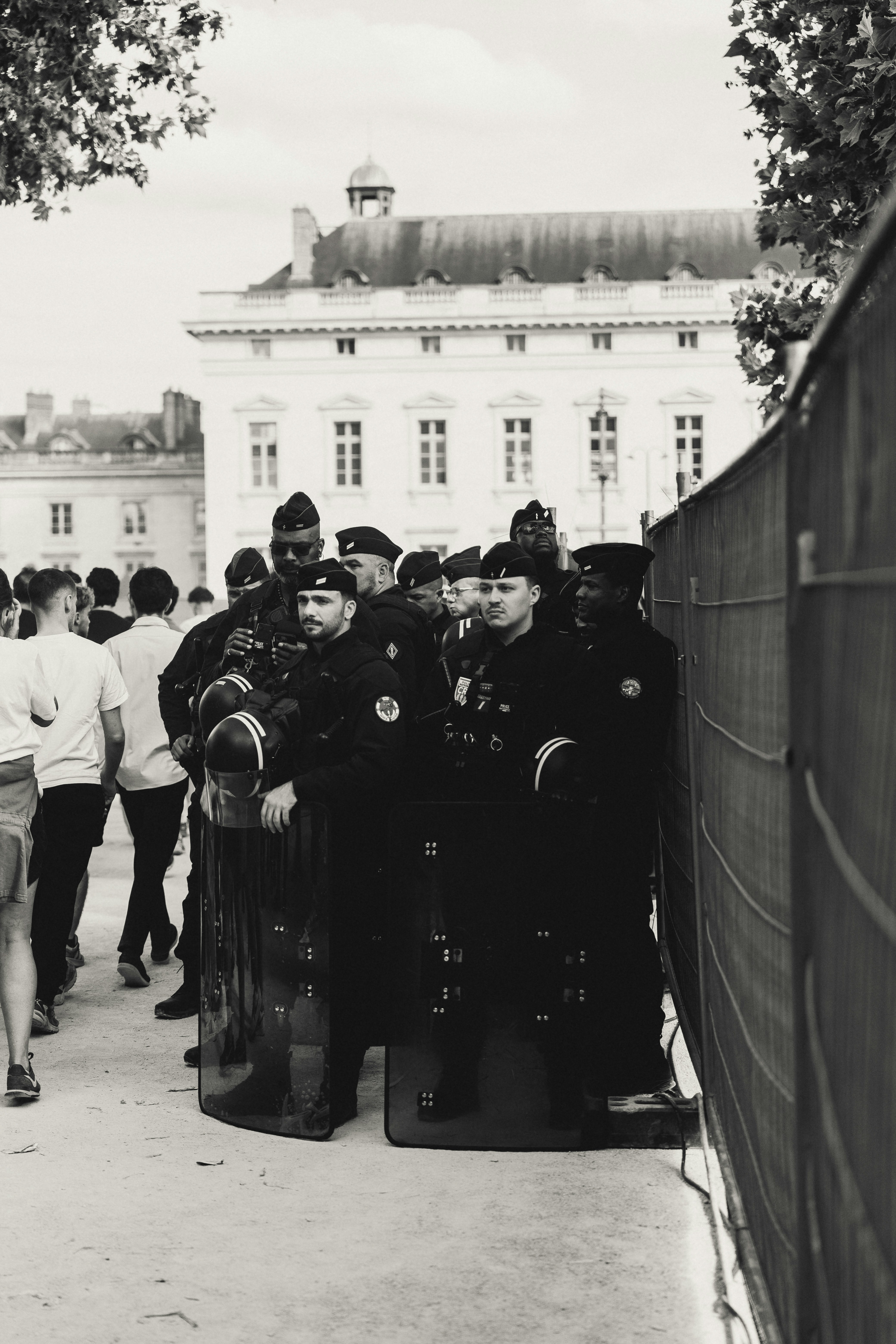 Police officers in uniform with shields stand by a fence.