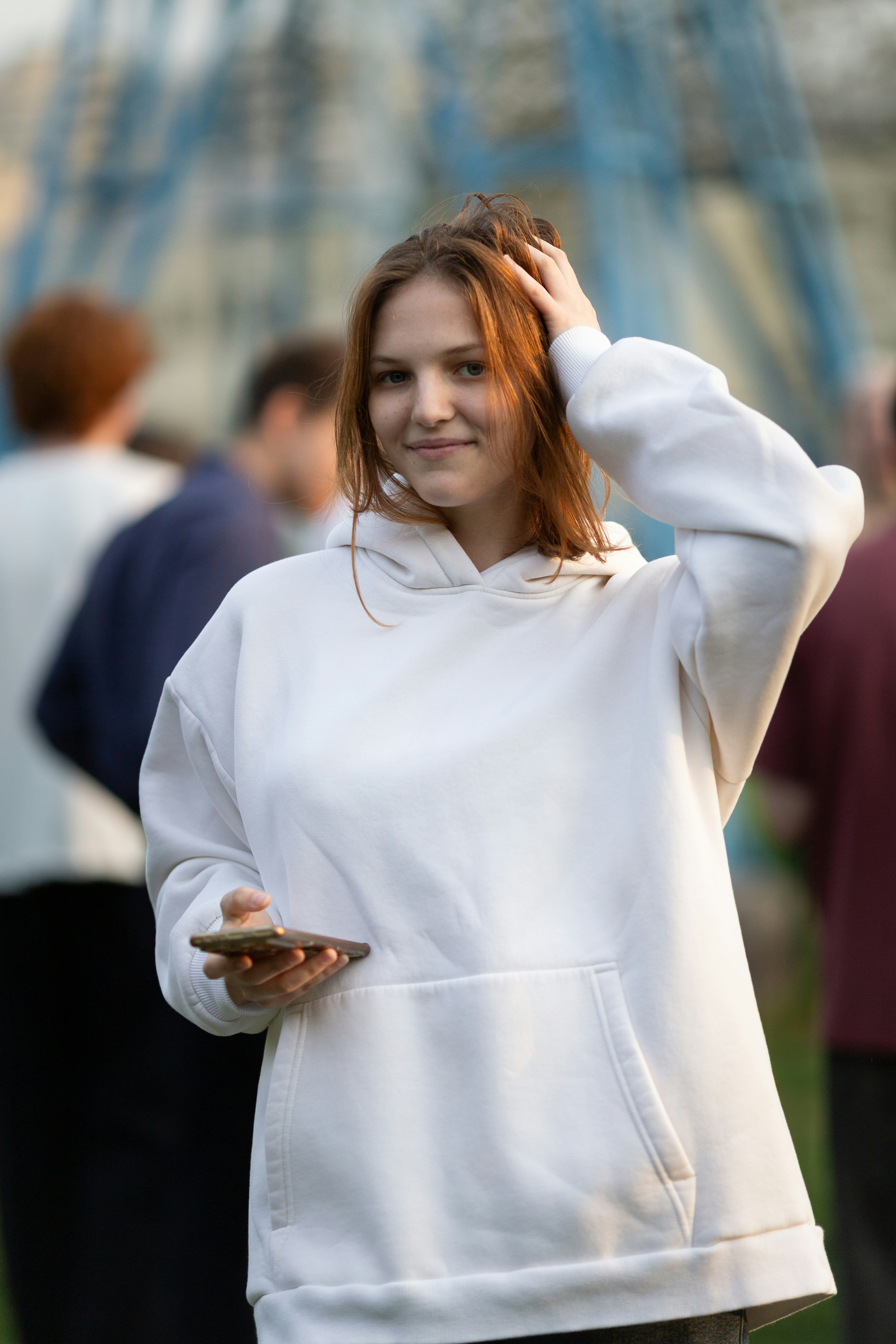 Smiling young woman in white hoodie holding phone outdoors.