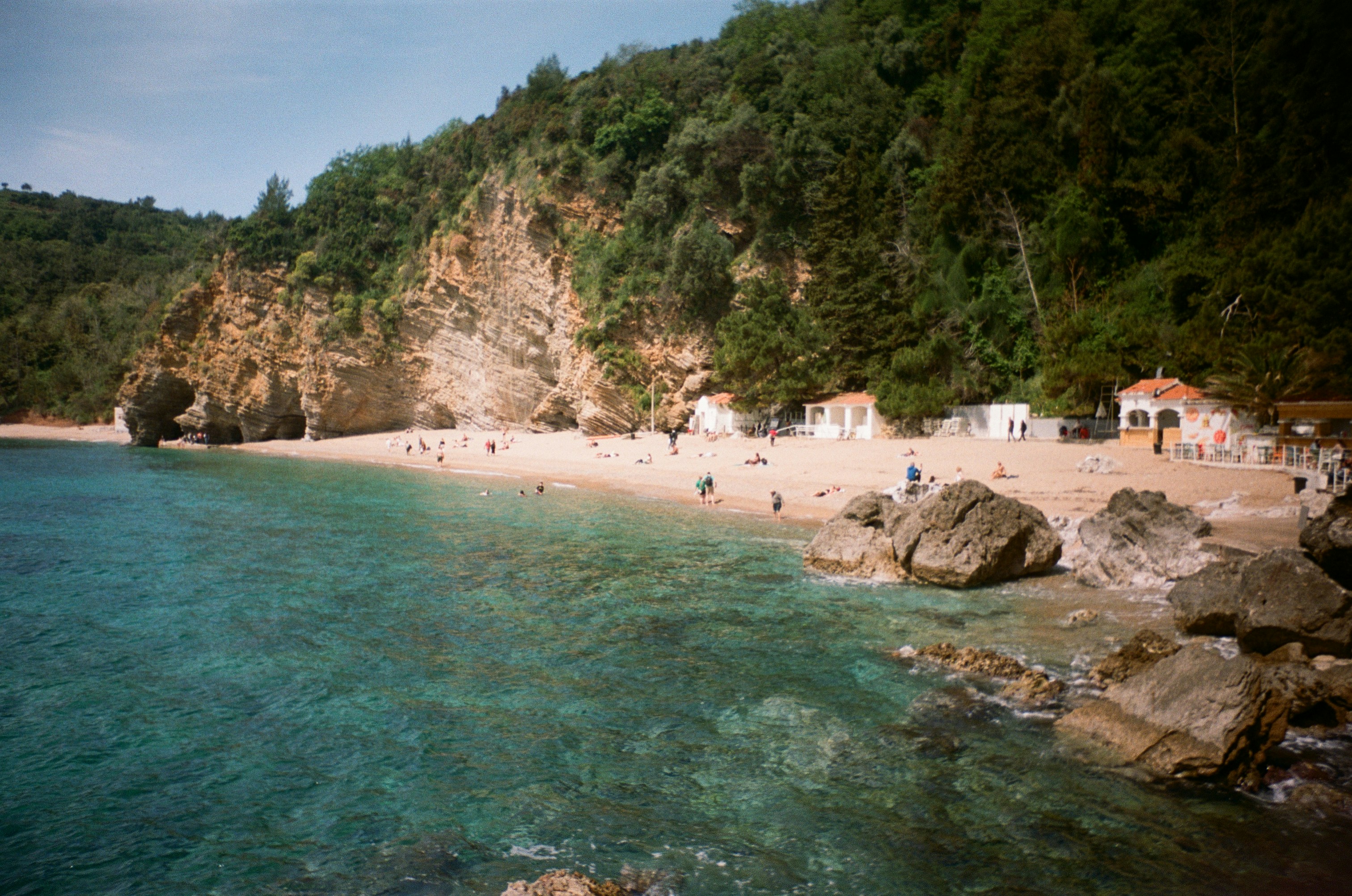 Sandy beach with clear turquoise water, cliffs, and green trees.
