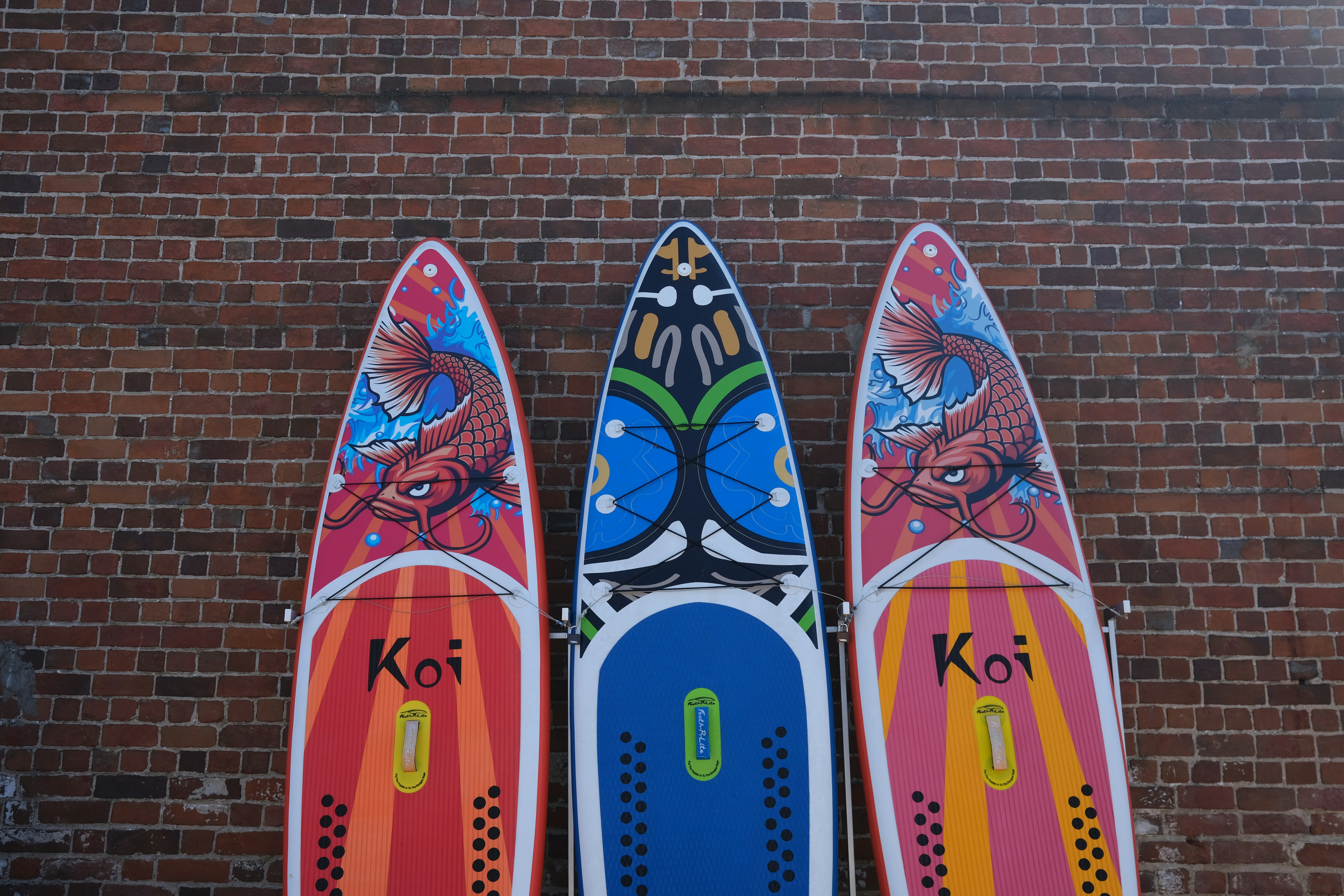 Three colorful paddleboards with designs leaning against a brick wall.