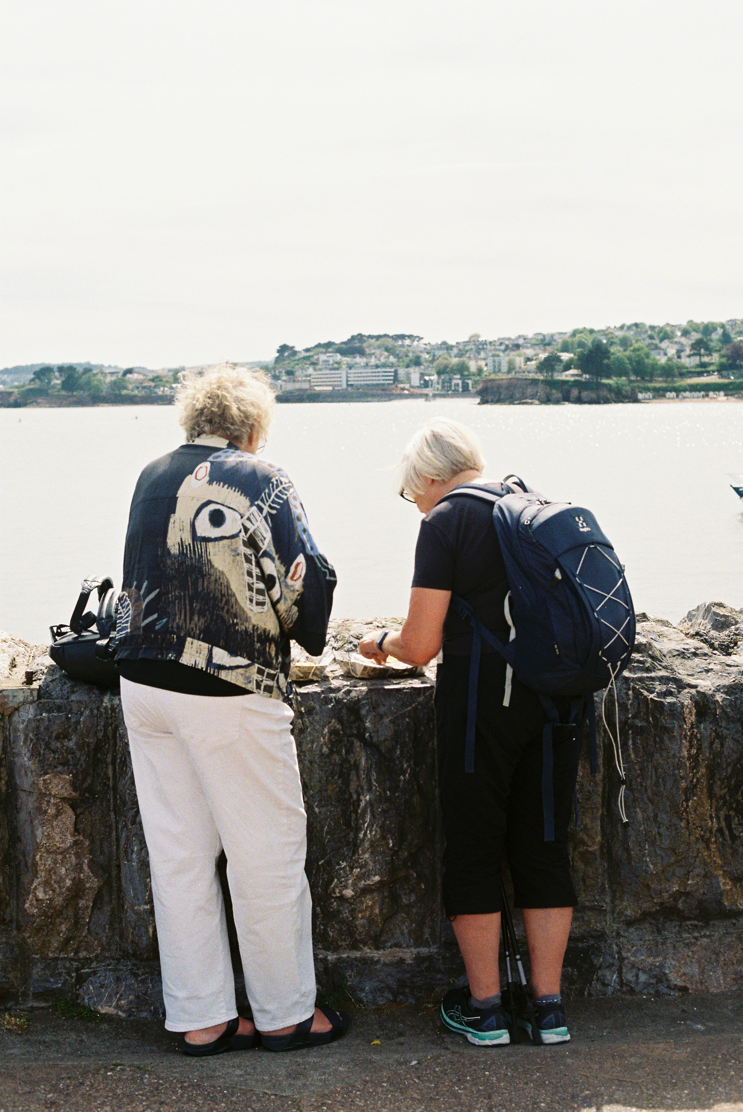 Women with backpack and artistic jacket view a coastal town.