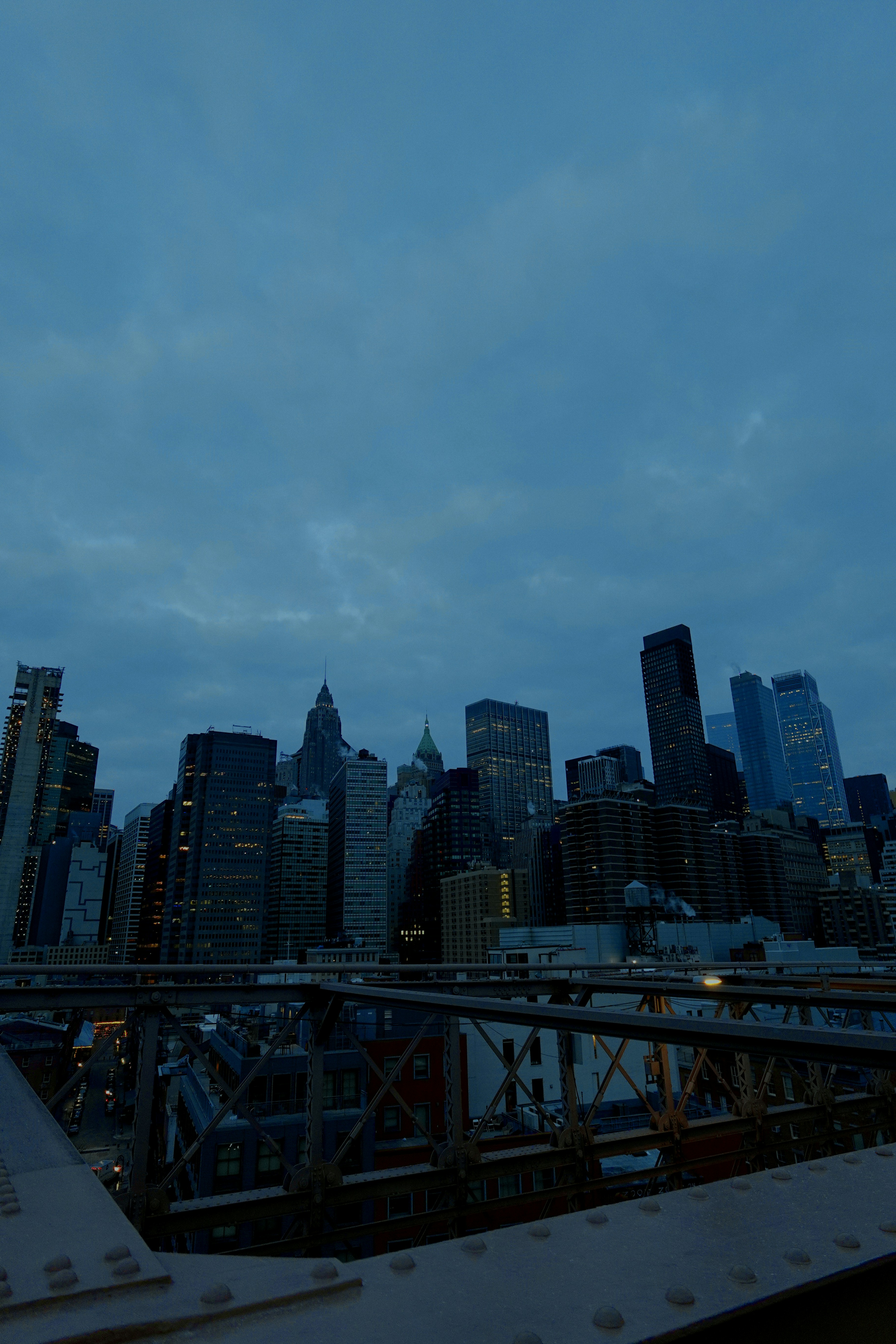 City skyline under dusky blue sky framed by a bridge railing and structures in foreground.