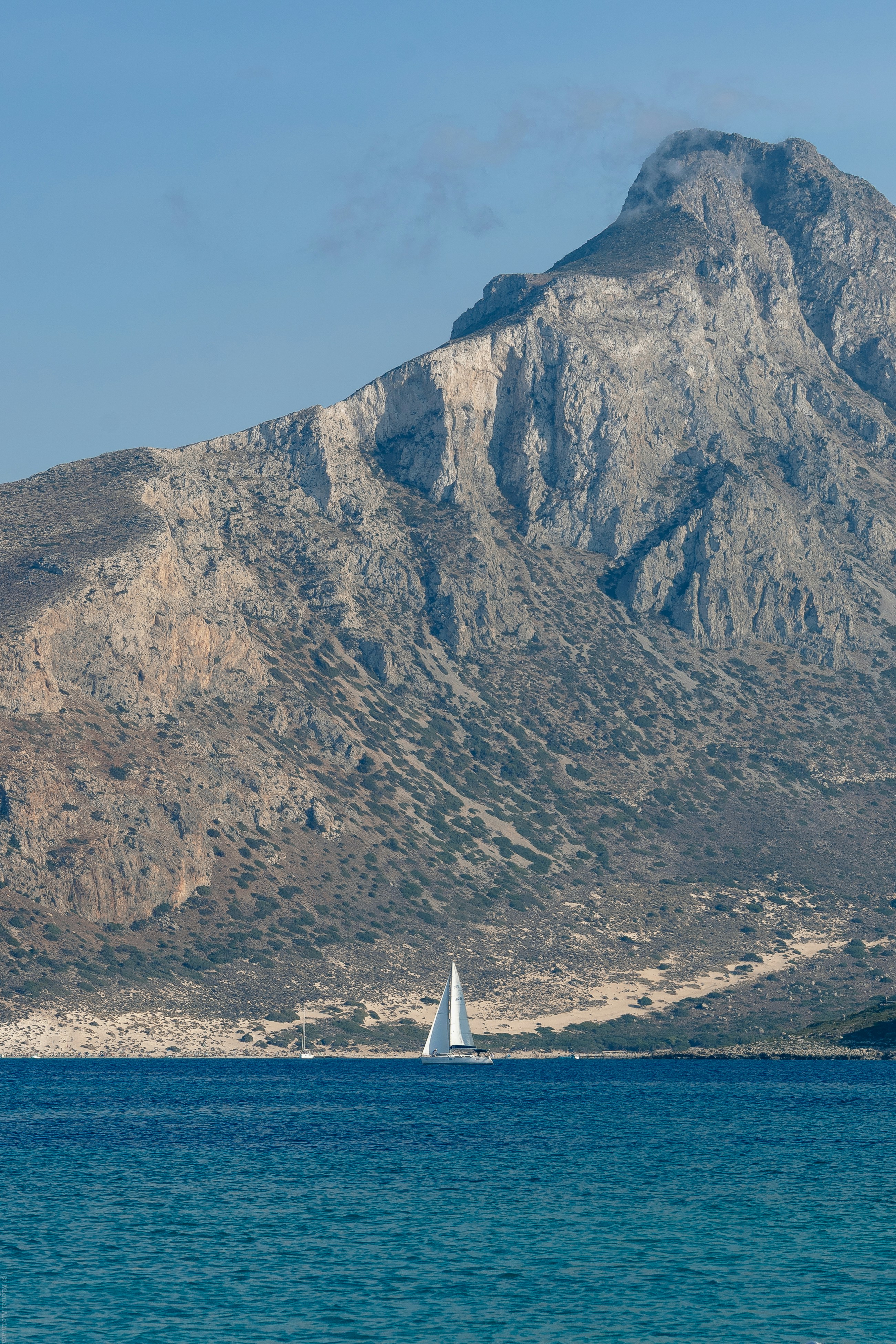 A sailboat on blue sea with a large rocky mountain.