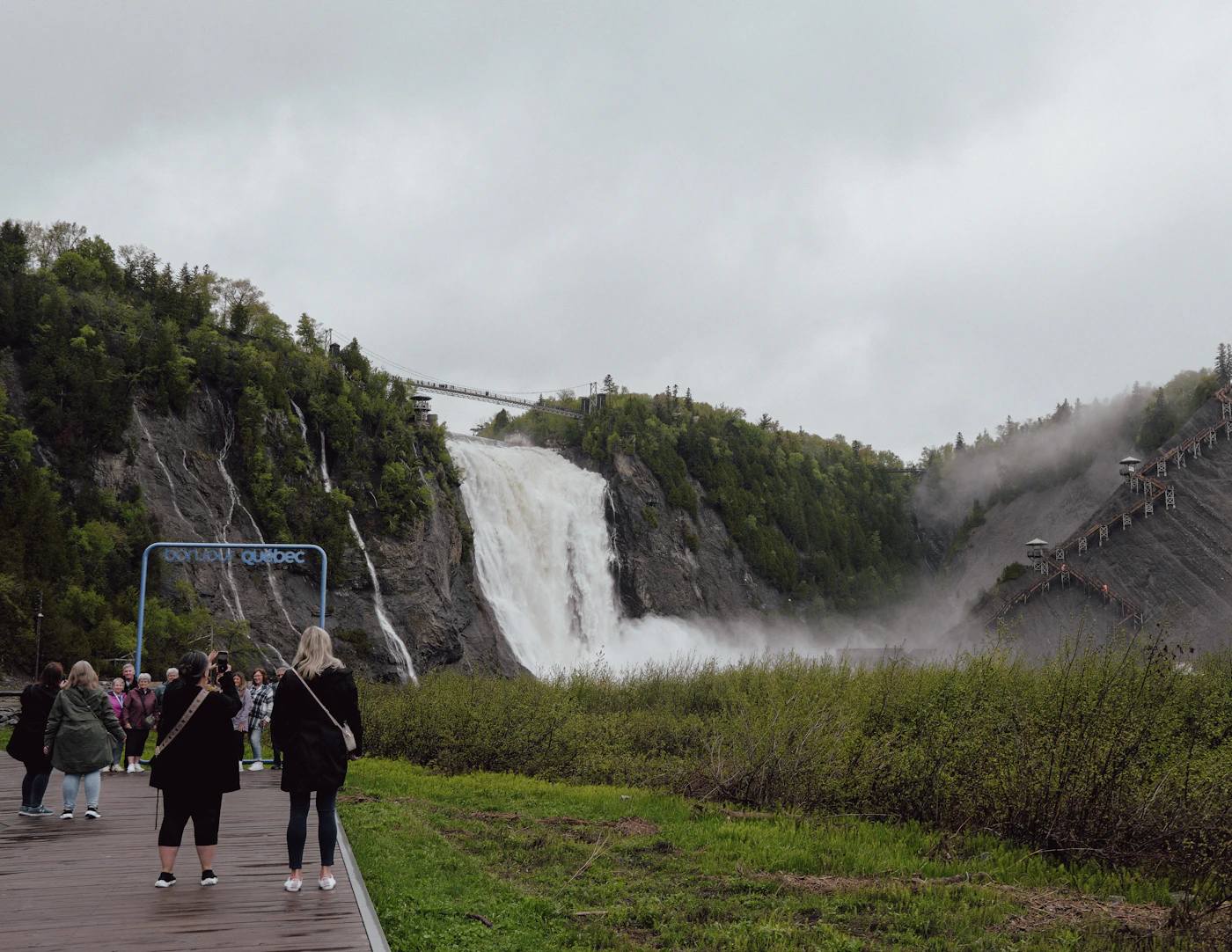 Group of tourists on a wooden viewing terrace observing the massive white cascade of Montmorency Falls near Quebec City with the dramatic waterfall spray visible