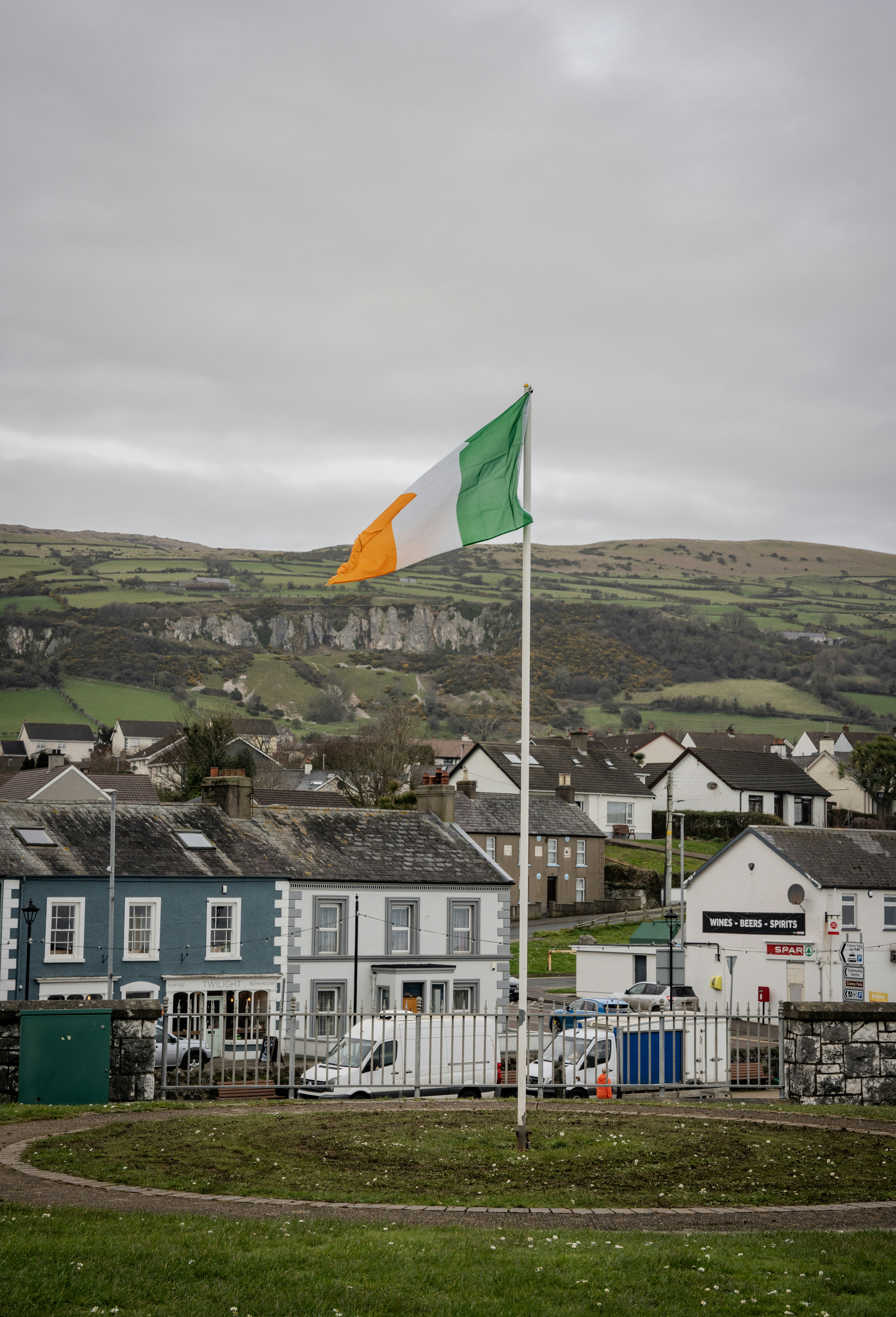 An irish flag flies over a village with green hills.