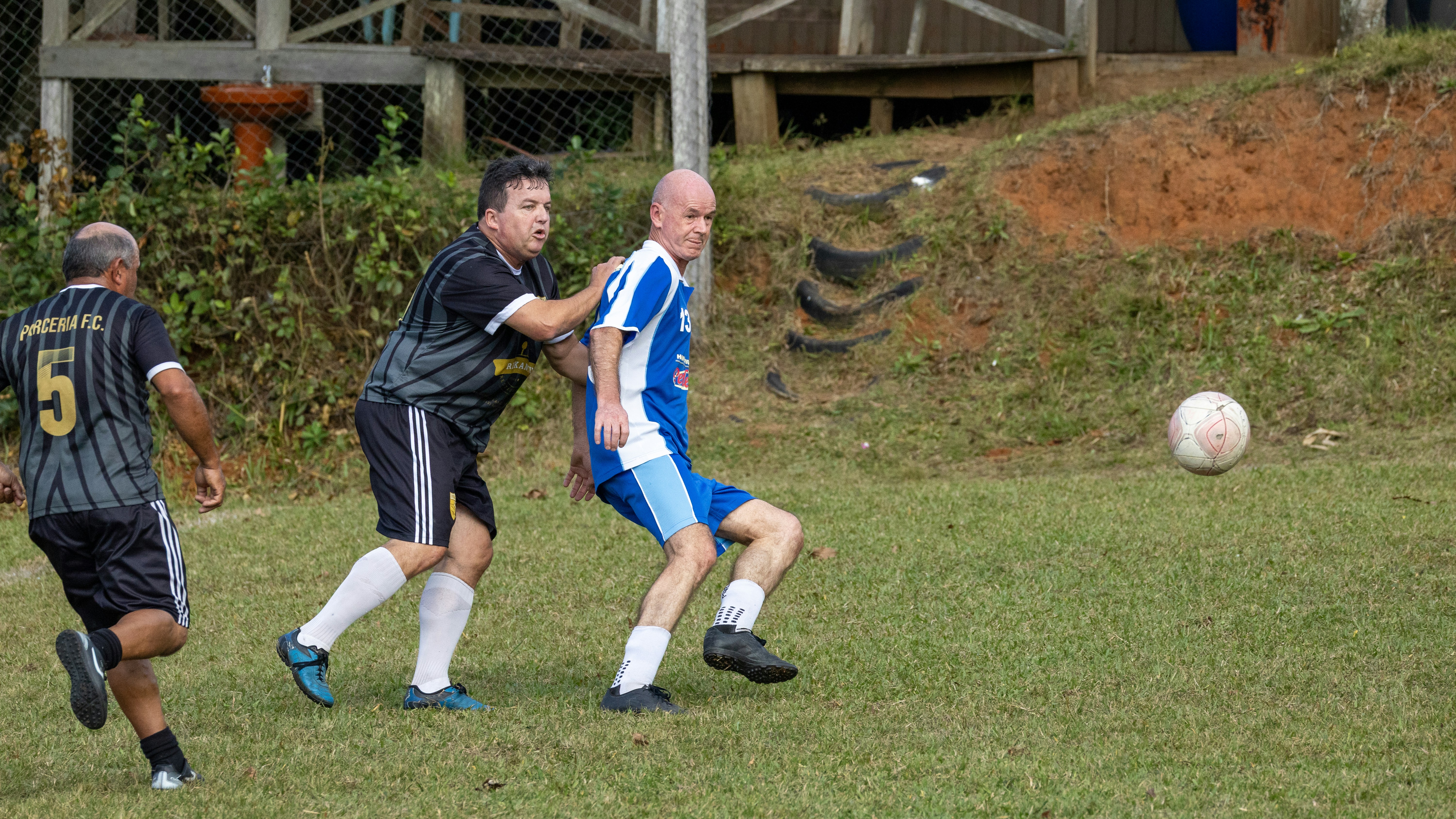Tiga pria bermain sepak bola di lapangan berumput.