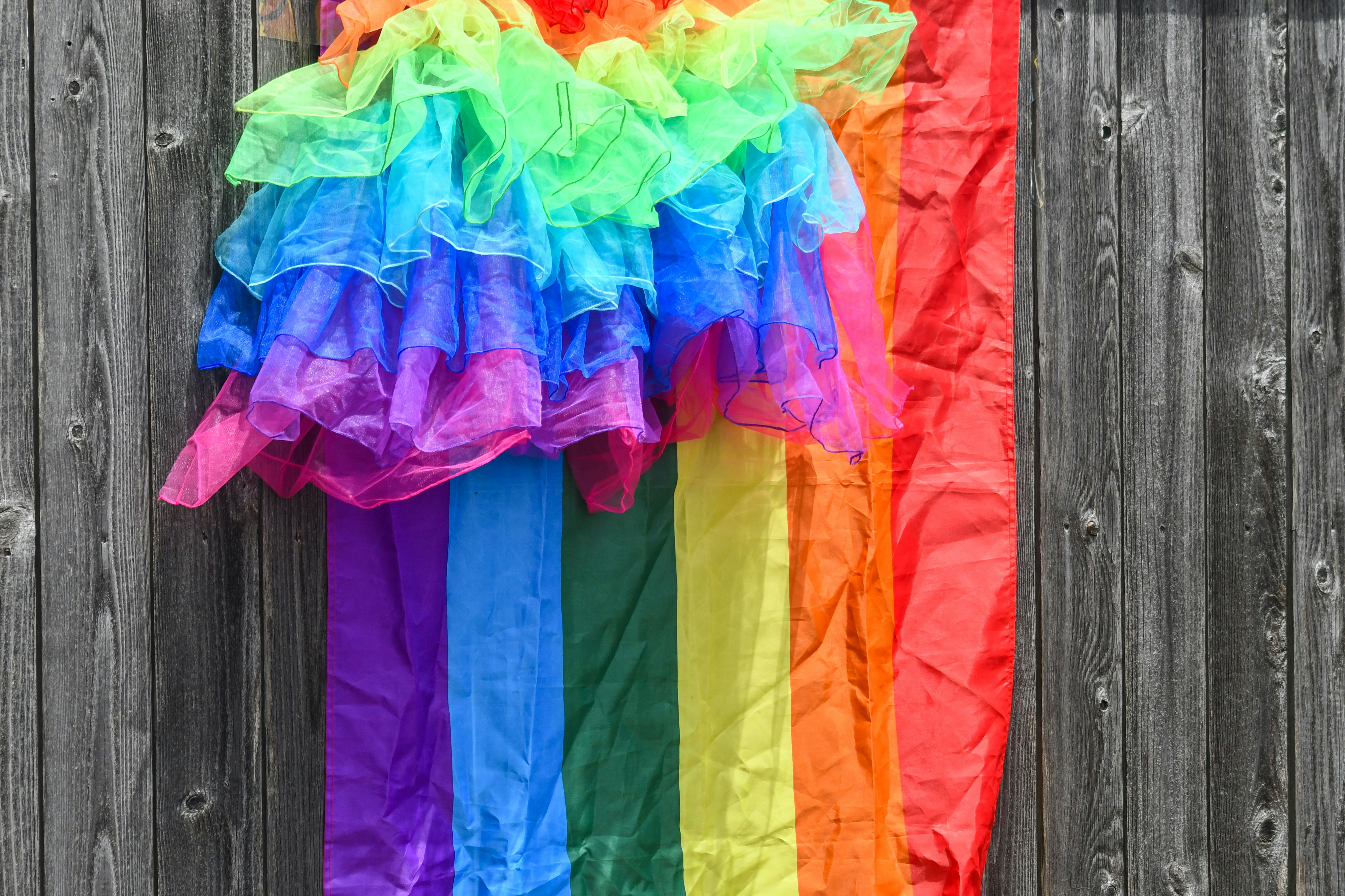 Vibrant rainbow fabric decoration hangs against a desaturated wooden fence.