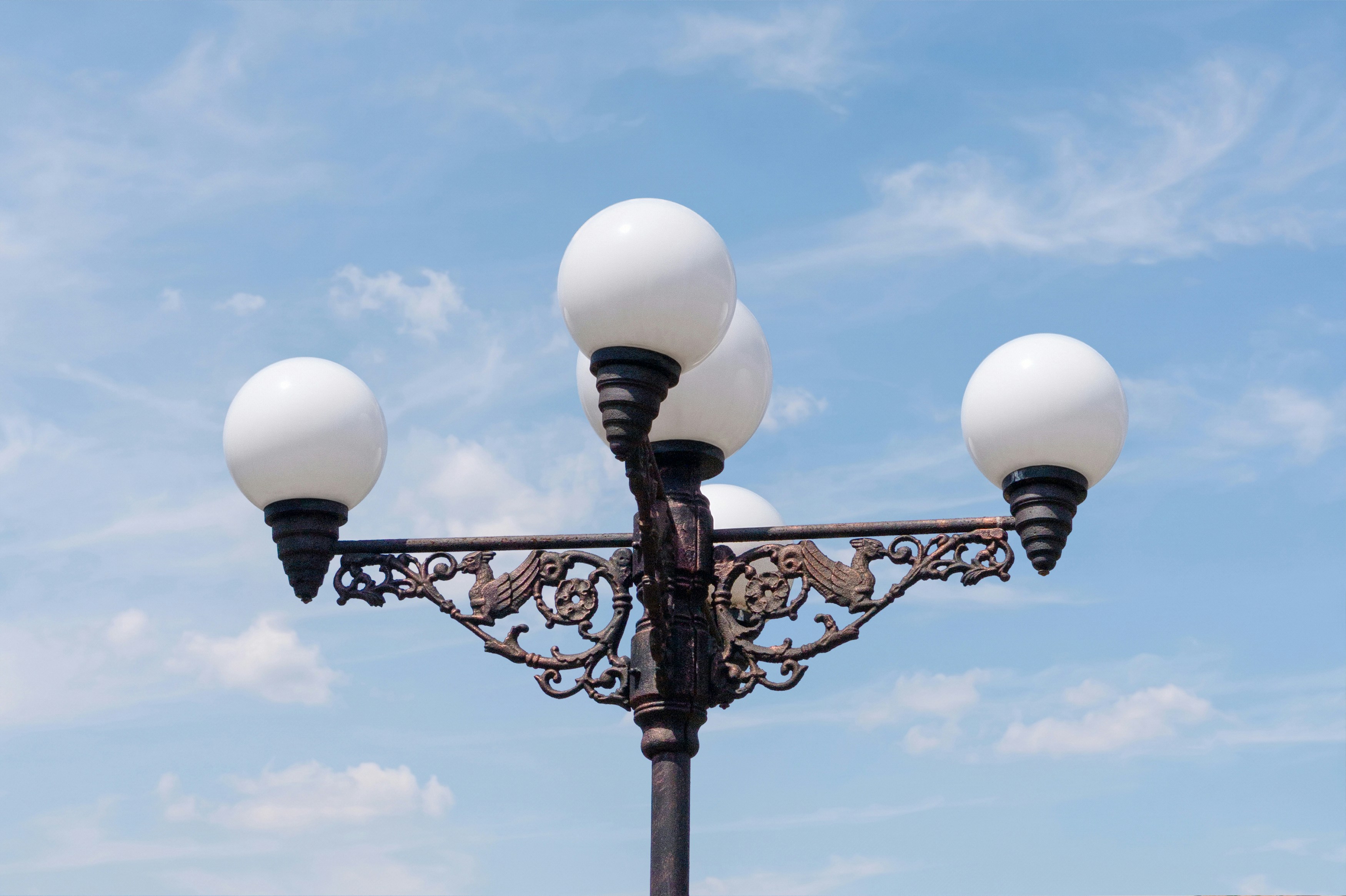 Verzierte Straßenlaterne mit vier kugelförmigen Lichtern vor blauem Himmel.