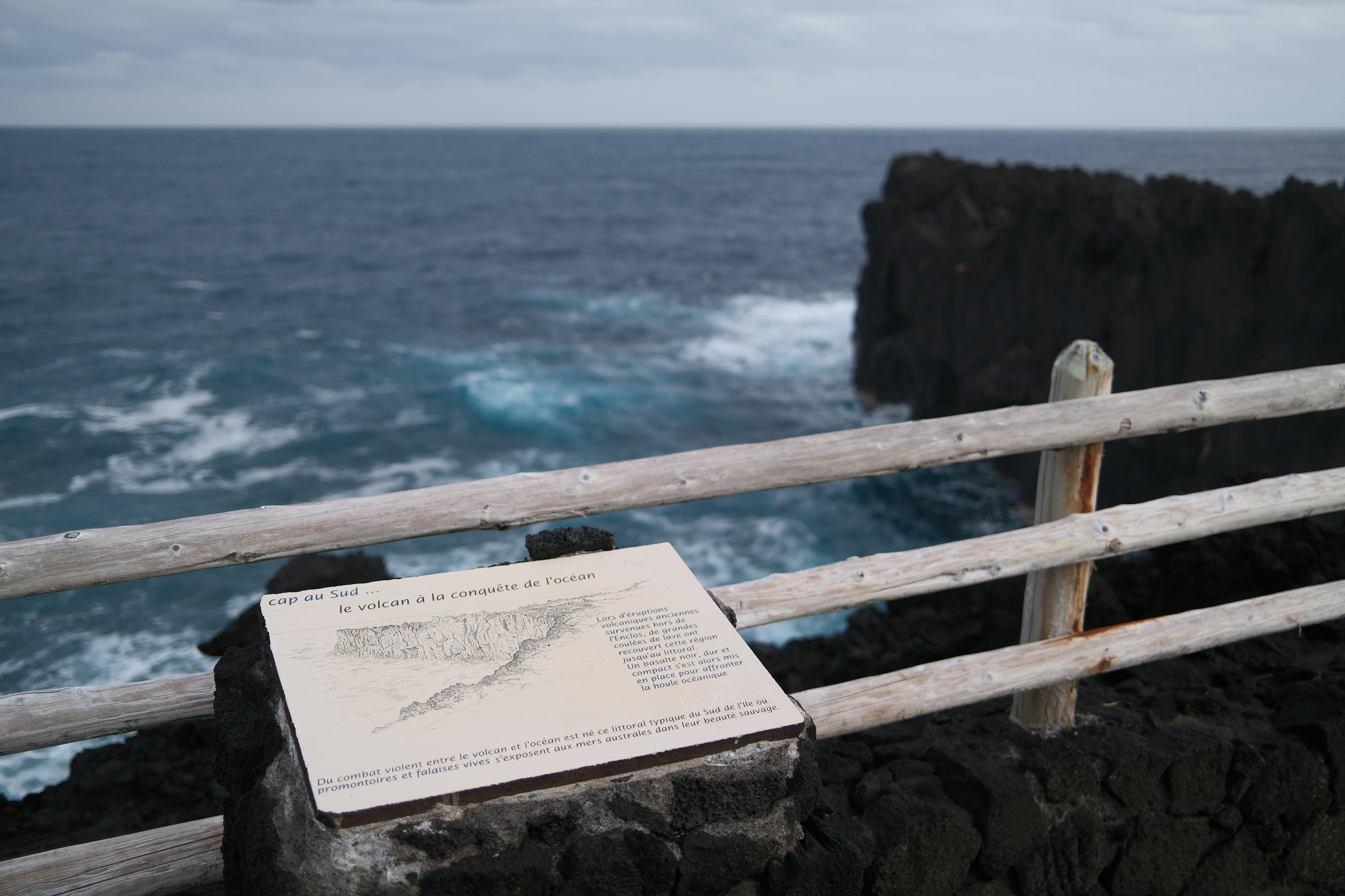 Information sign by a wooden fence overlooking a volcanic coast.