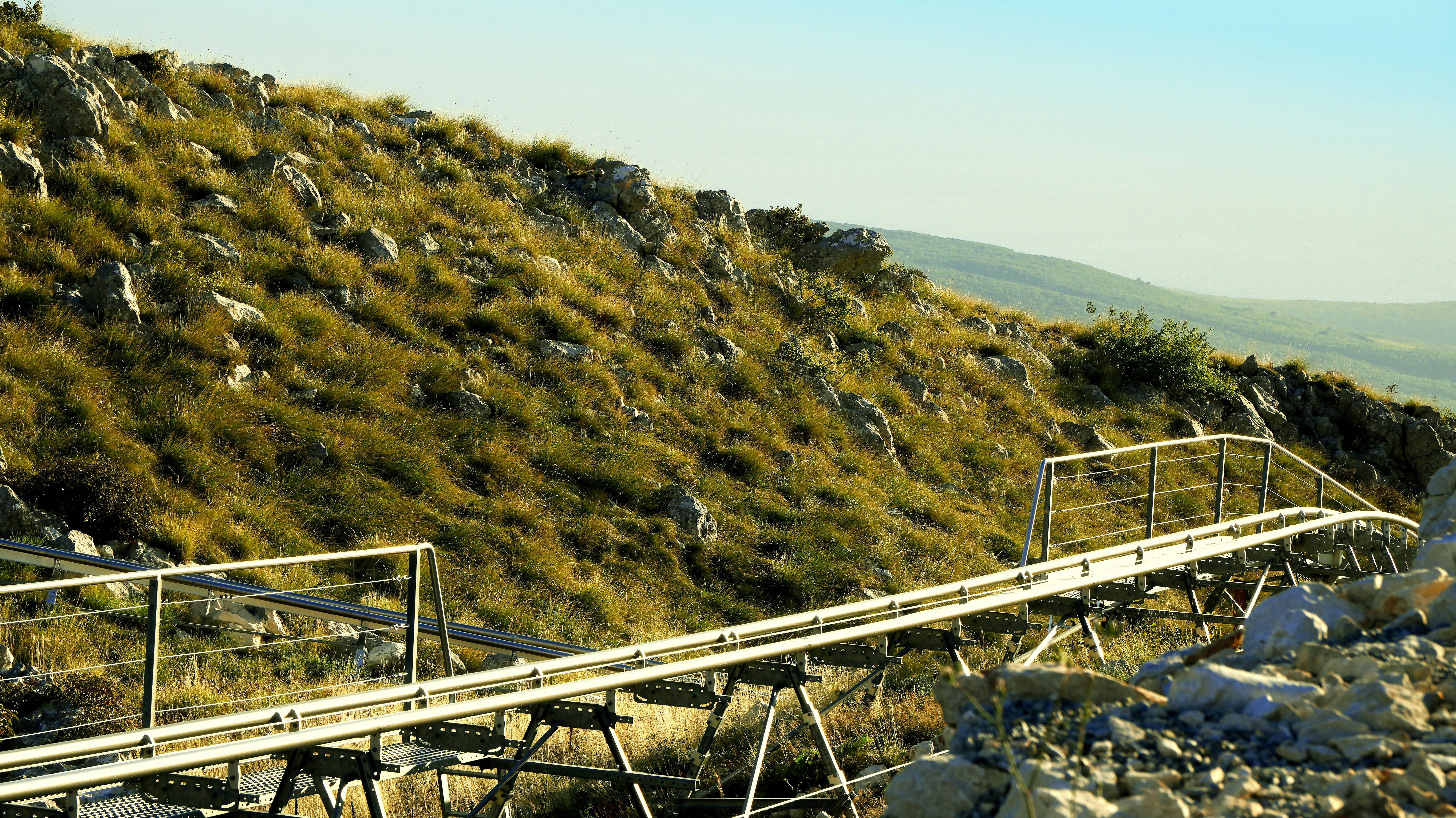 A metal walkway extends along a steep, grassy mountain slope.