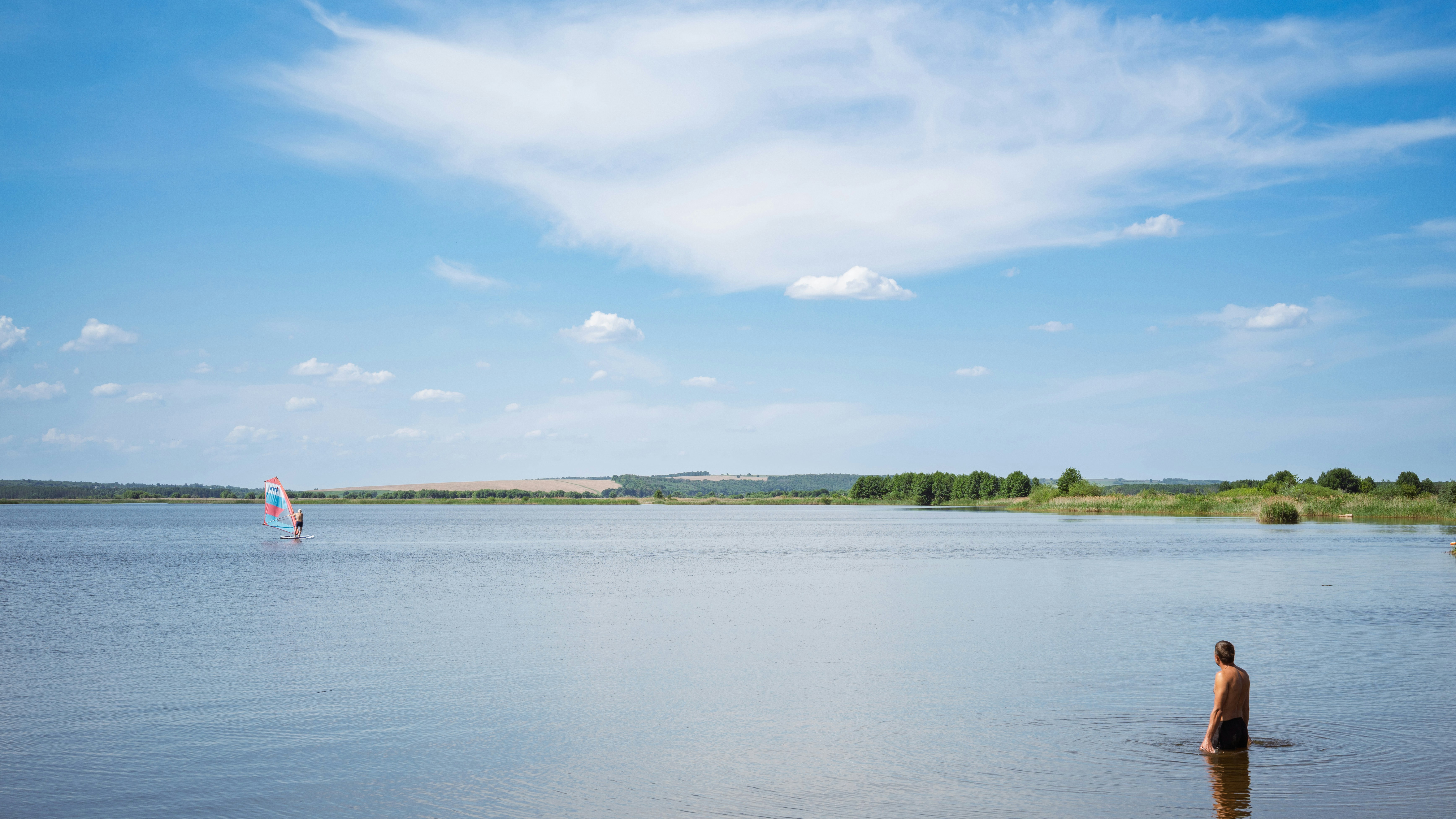 A man wades in a calm lake with a windsurfer.