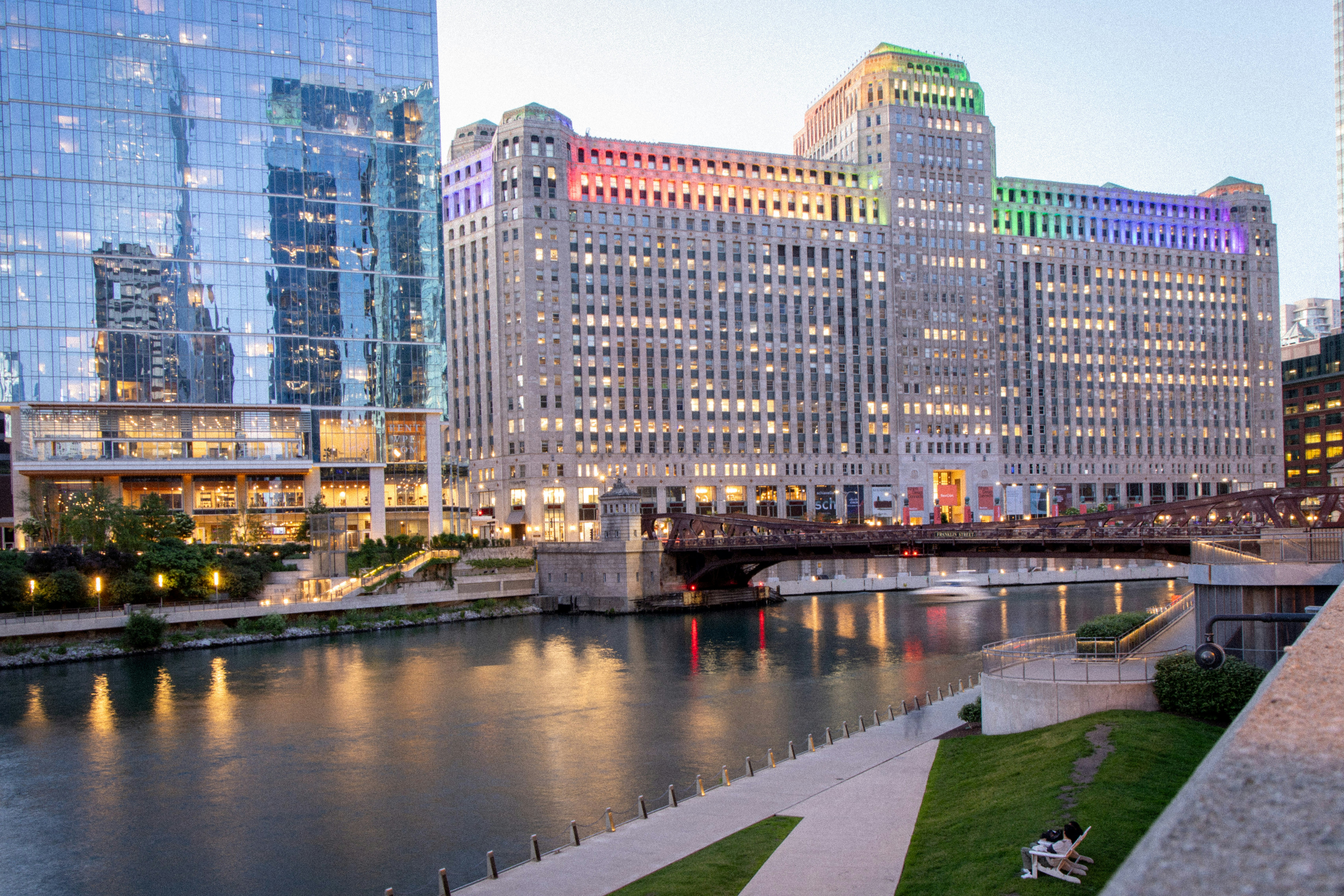 Building with rainbow lights reflects in city river at dusk.